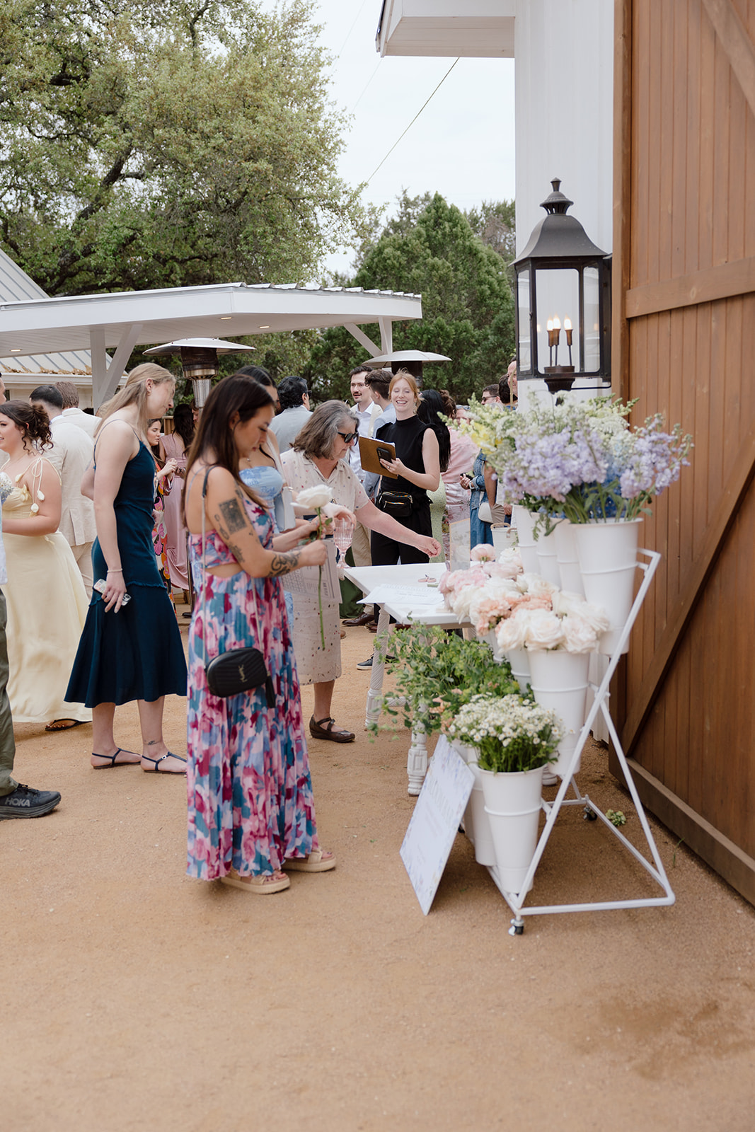 Group of people gathered outdoors near a table with flowers and lanterns, trees and a building in the background.