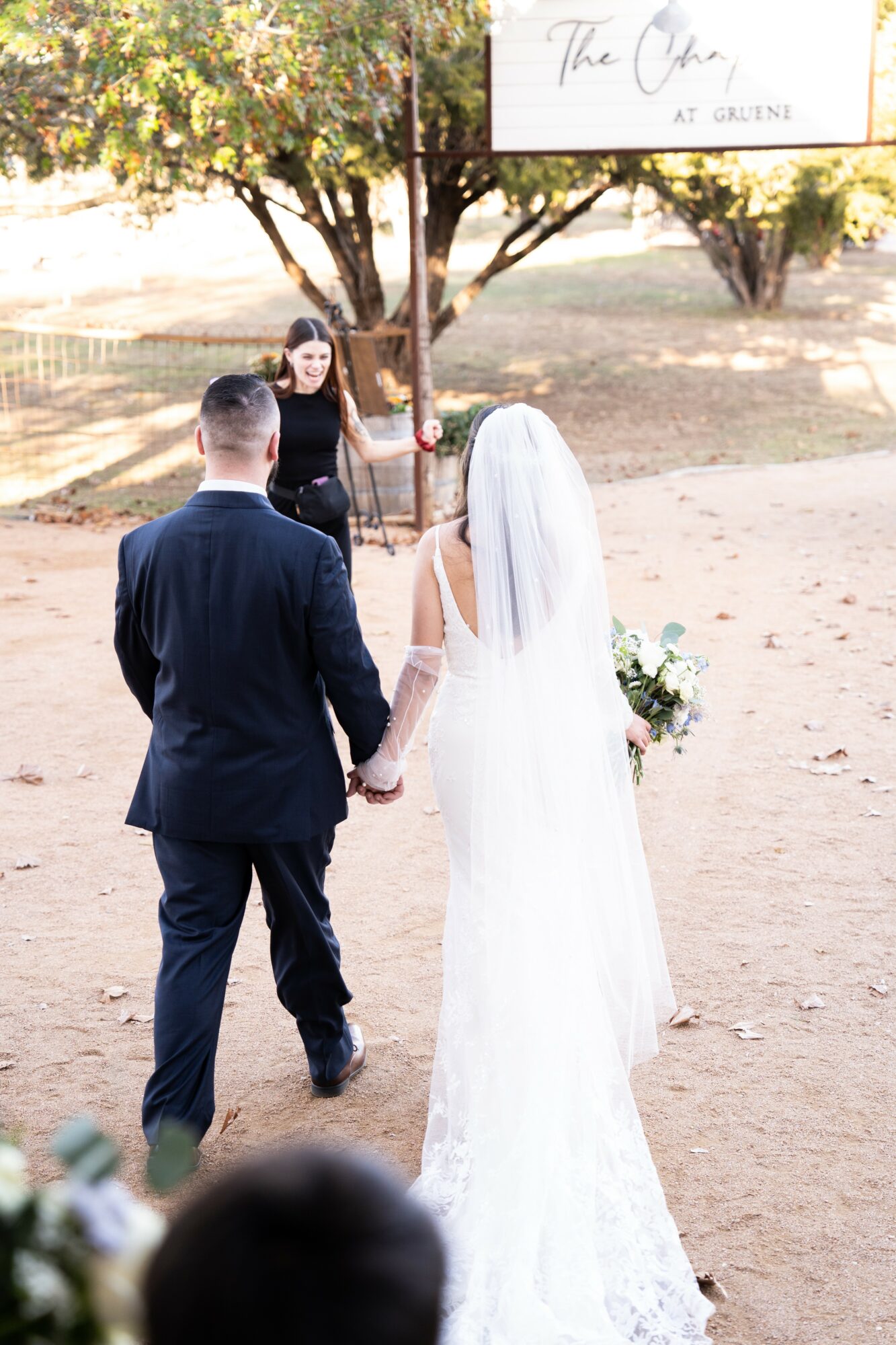 Bride and groom holding hands facing an officiant outdoors, with trees and a sign in the background.