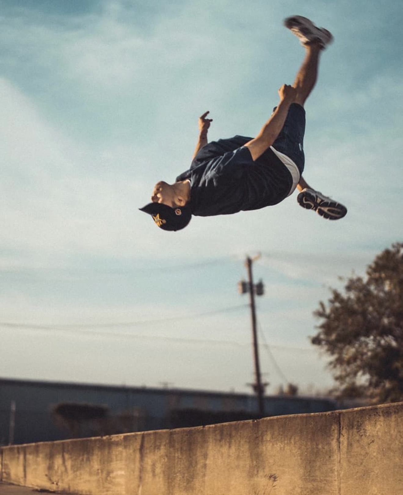 Person mid-air performing a flip outdoors, sky and trees in background, wearing dark clothing and cap.