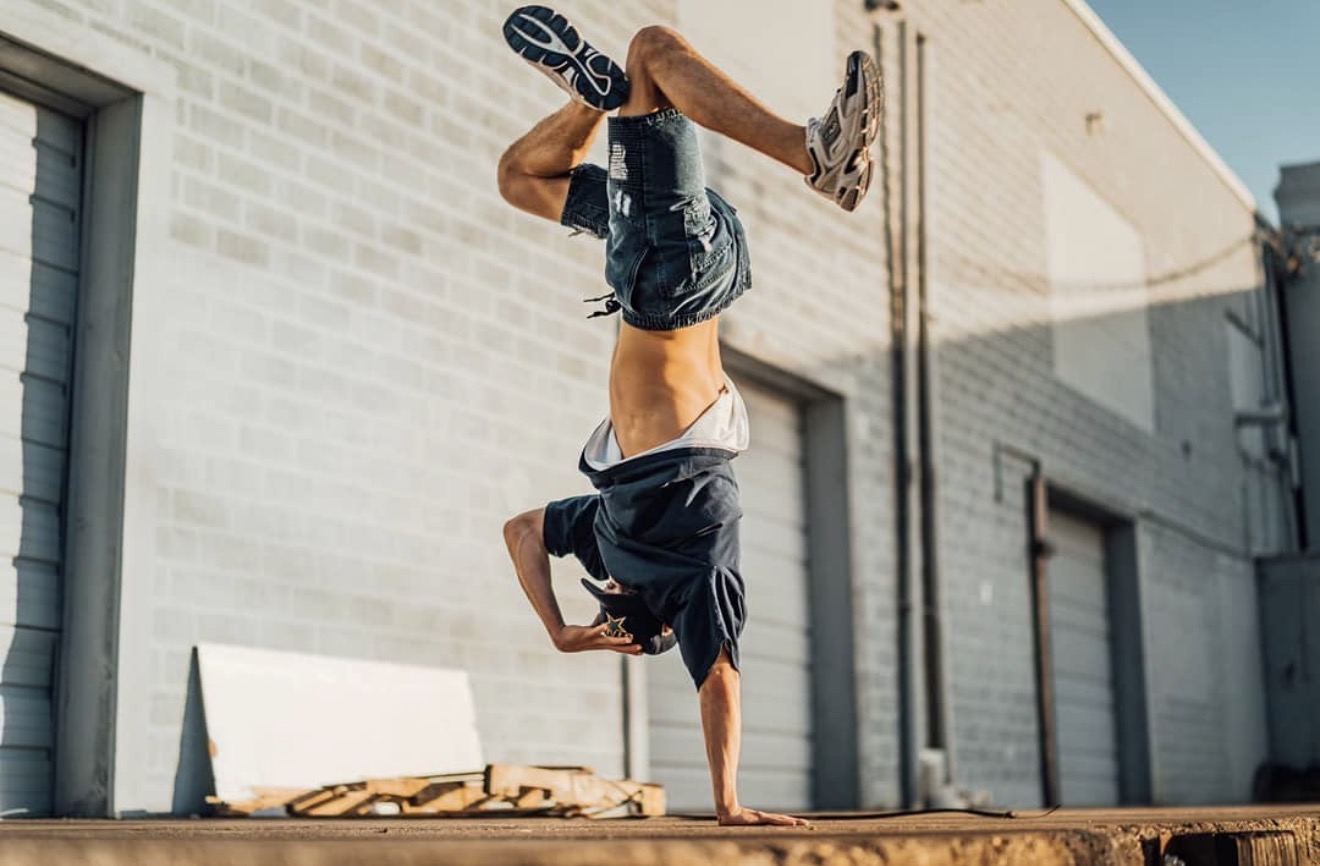 Person performing a handstand against a wall outdoors, with legs bent and head near the ground.