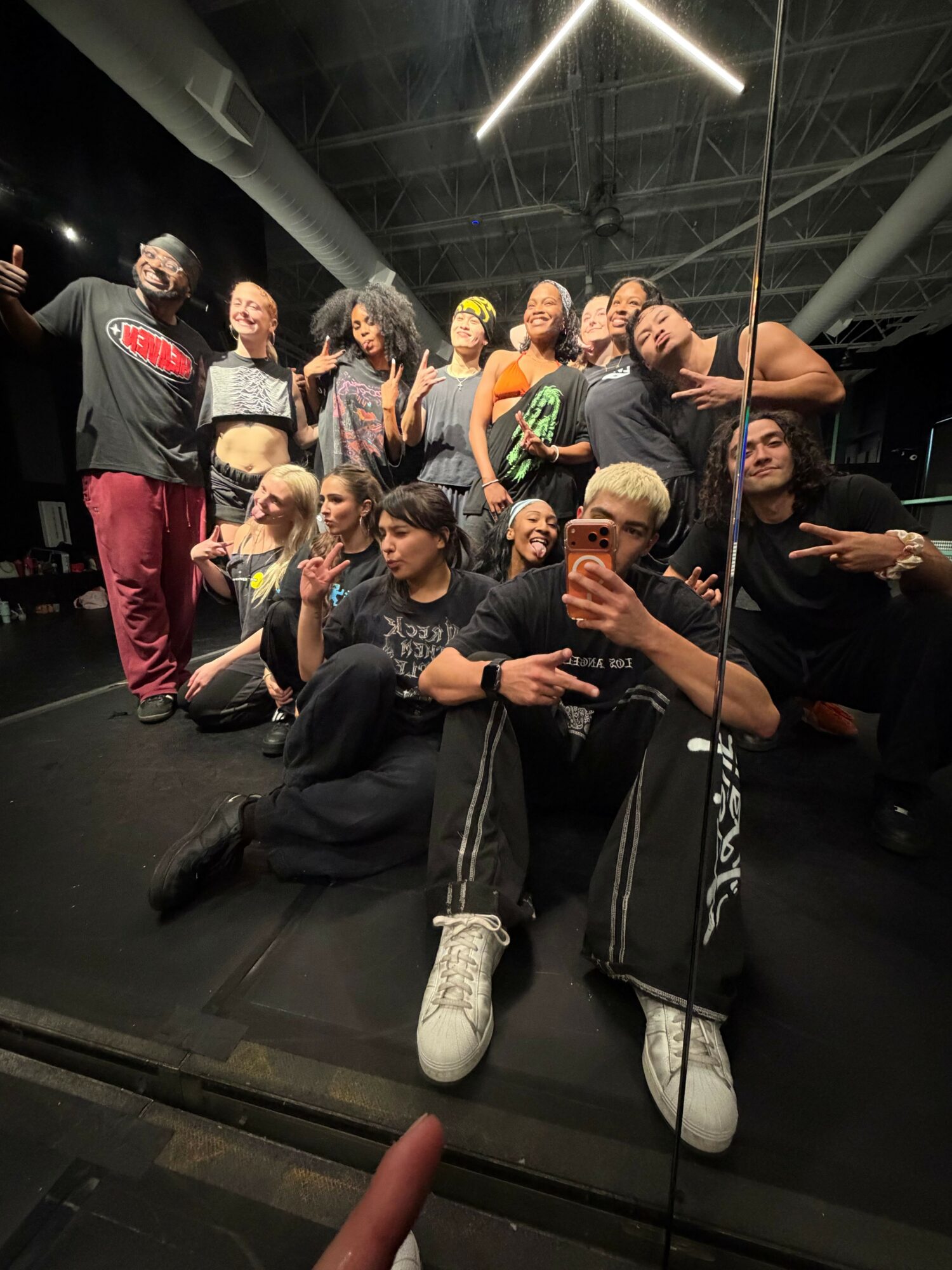 Group of people taking a selfie in a mirror, some making peace signs, in a dark indoor space.
