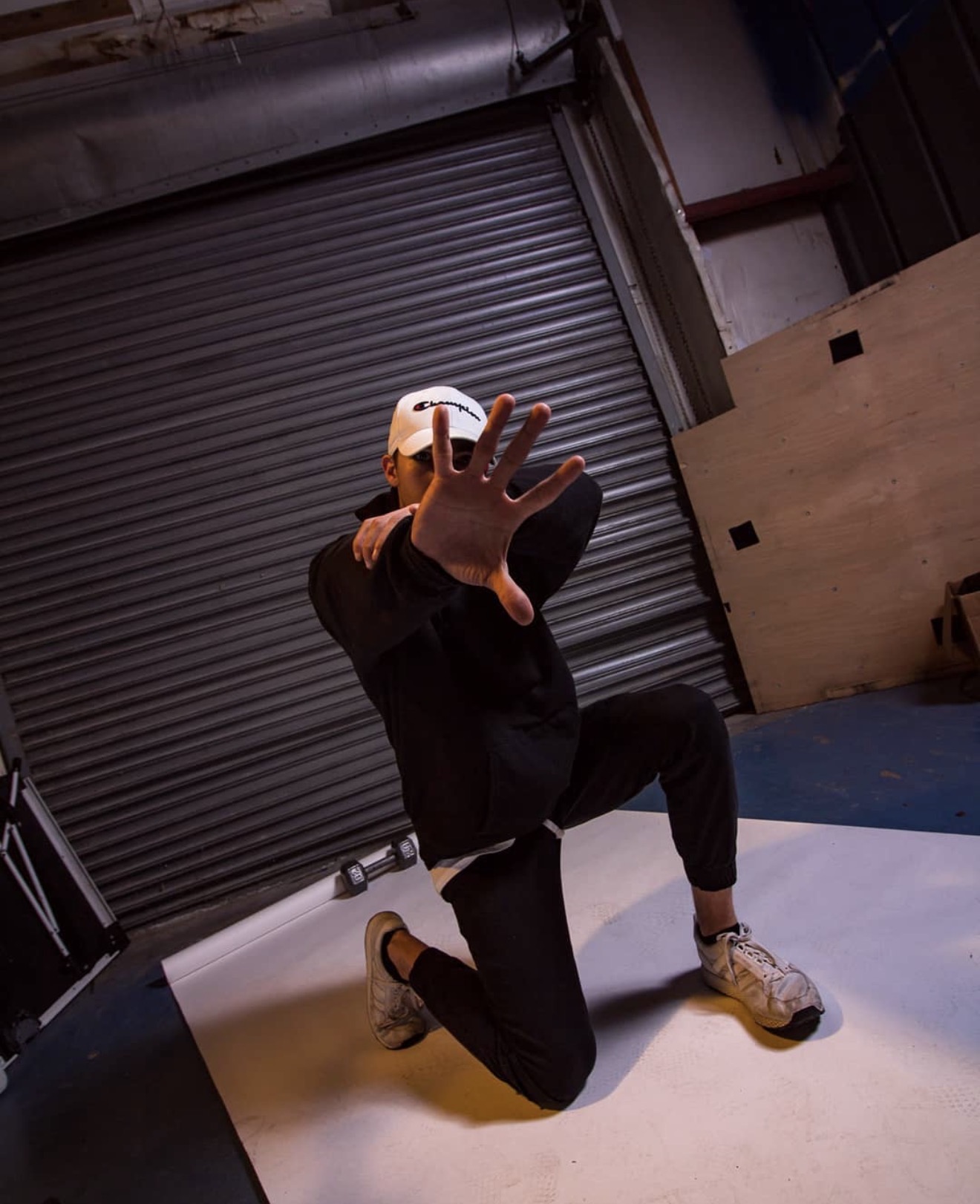 Person kneeling indoors, reaching toward the camera with hand extended, wearing a cap, dark clothing, and sneakers.