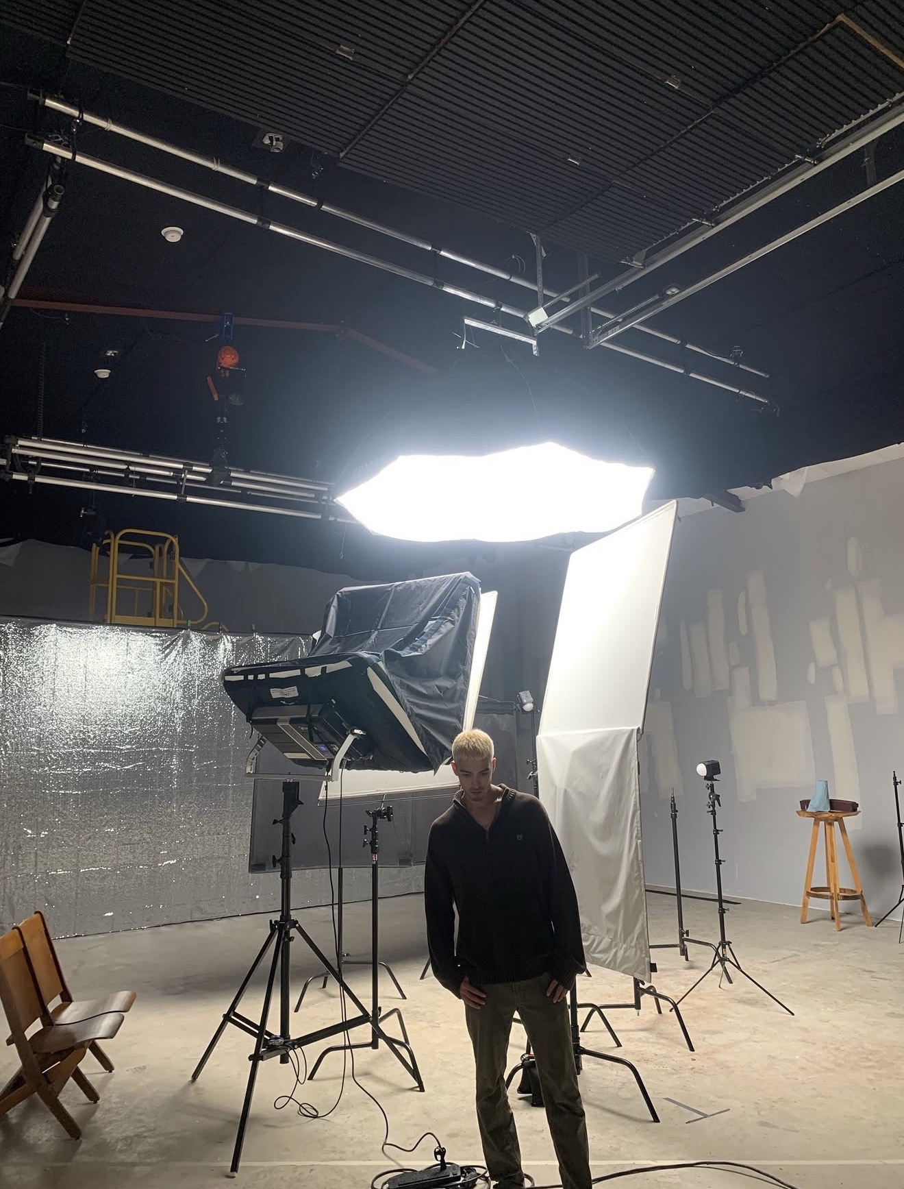 Person standing in a photography studio with lighting equipment and a white backdrop, ceiling with metal beams, and a stool.