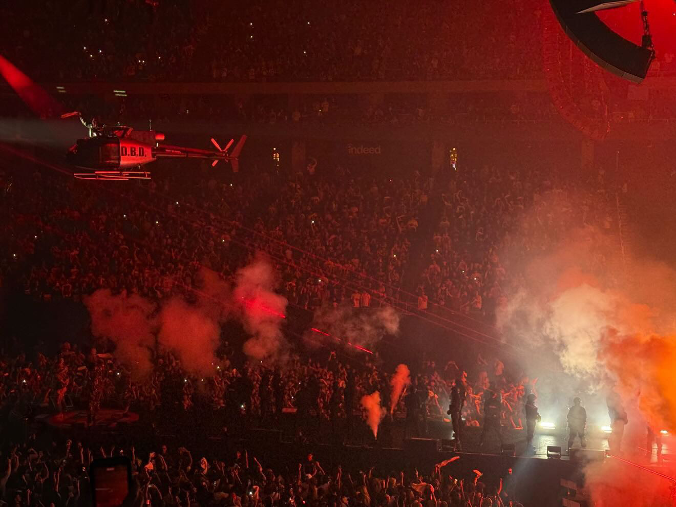 Large crowd at concert with red lighting, smoke effects, and stage in background.