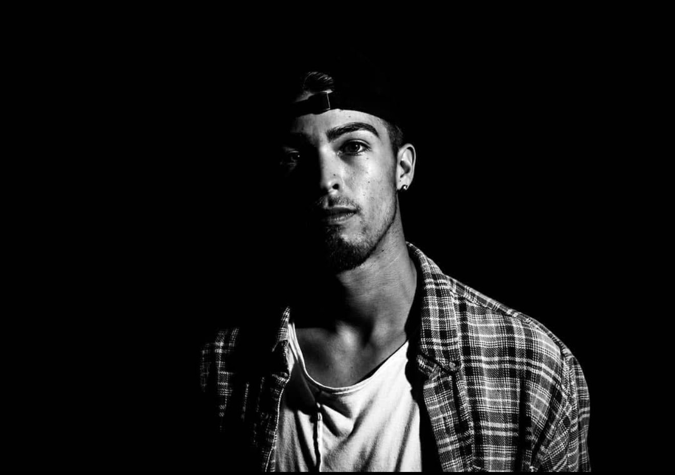 Young man with a cap, plaid shirt, and white t-shirt, looking at the camera against a black background.