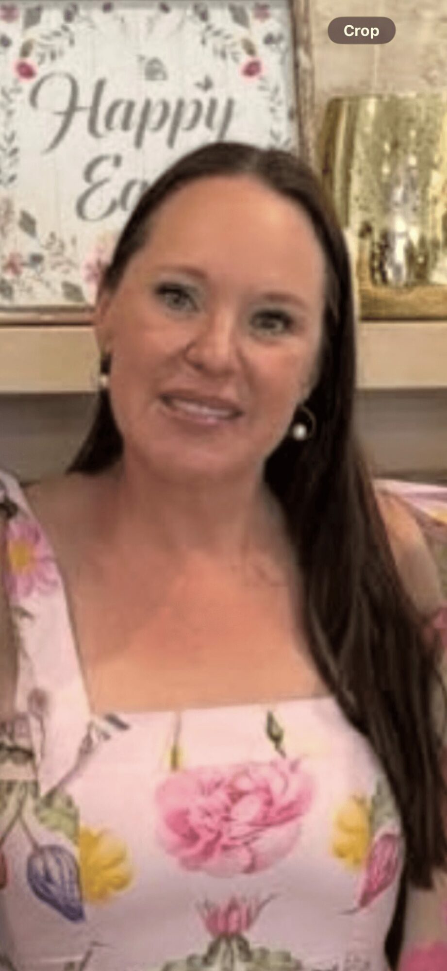 Woman with long dark hair smiling, wearing a floral dress, in front of a festive background with a sign and gold decorations.