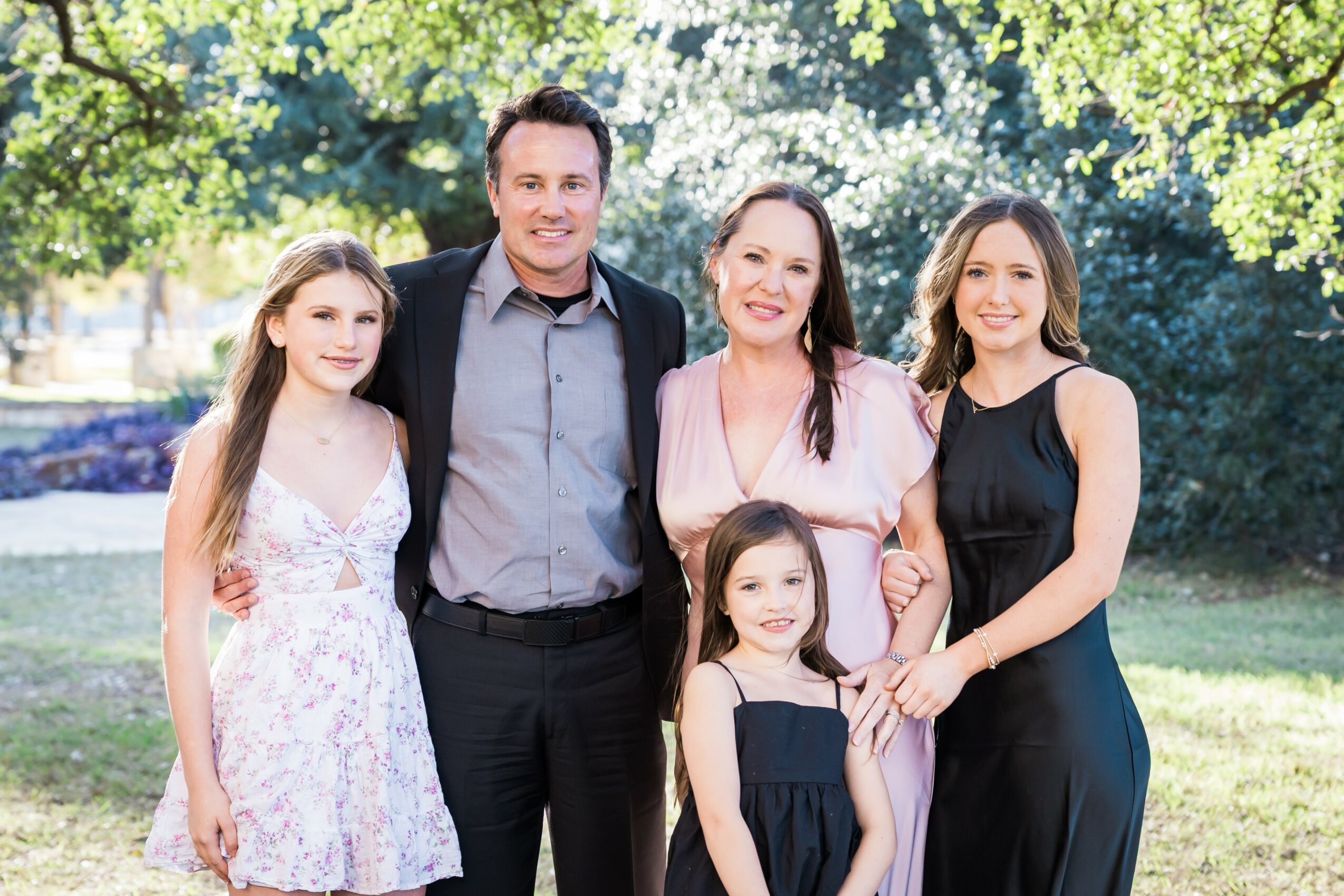 Family of five outdoors, smiling, standing on grass with trees in background, two adults and three children, all dressed nicely.