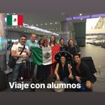 Group of people holding a Mexican flag at an airport, smiling, with a sign that says 'Viaje con alumnos'.