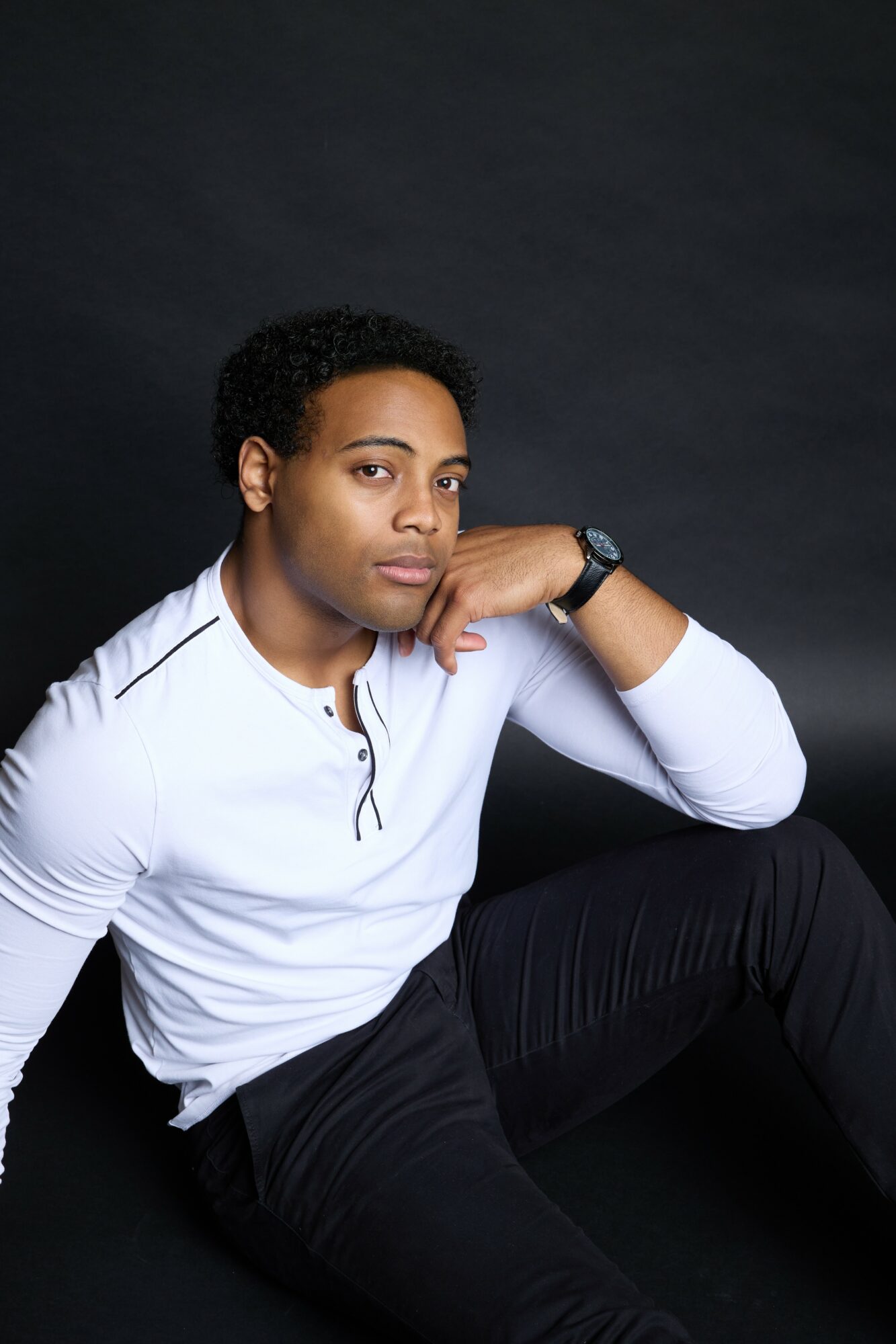 Person with short curly hair wearing a white long-sleeve shirt and black pants, sitting against a dark background.