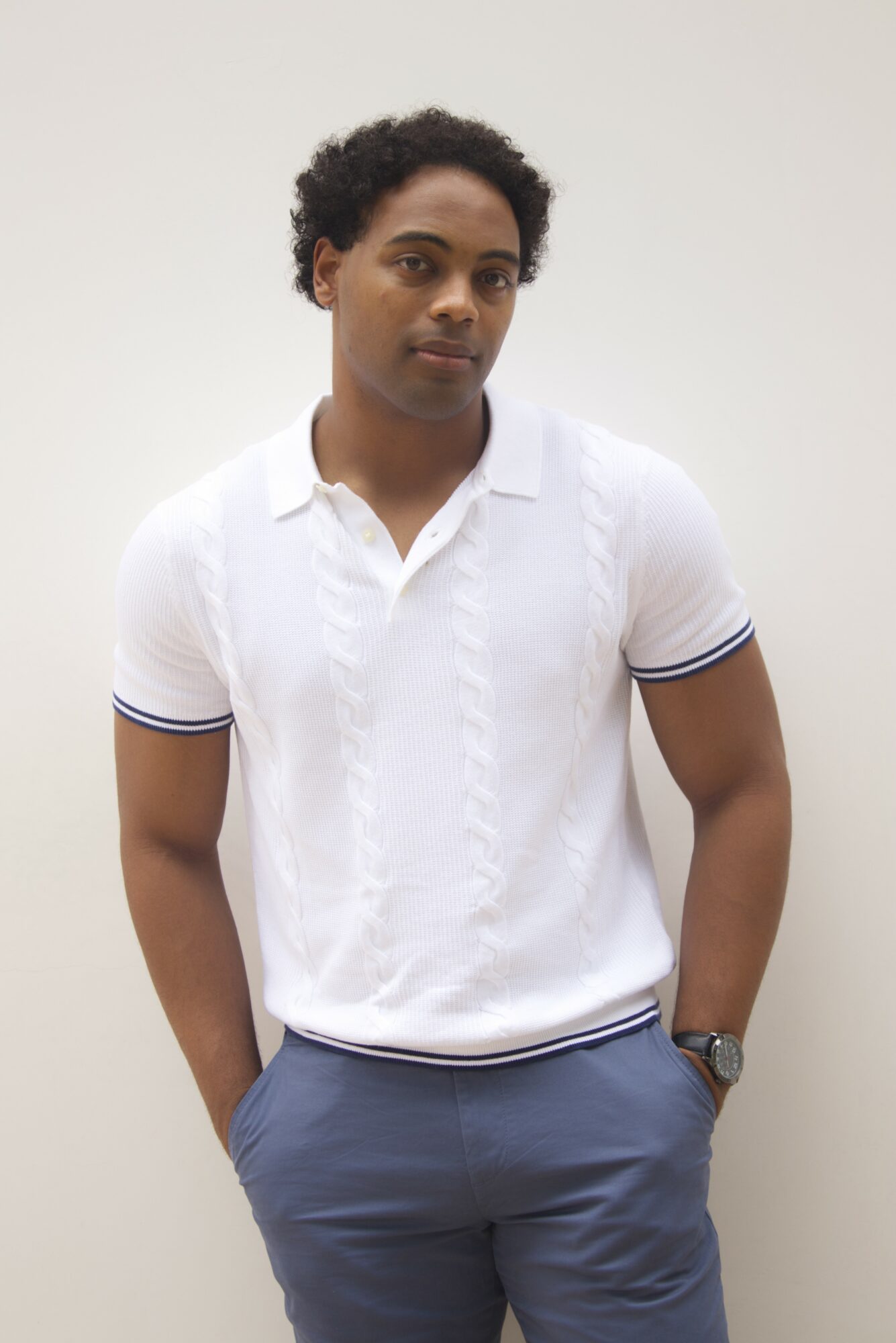 Young man with curly hair wearing a white polo shirt and blue pants, standing against a plain background.
