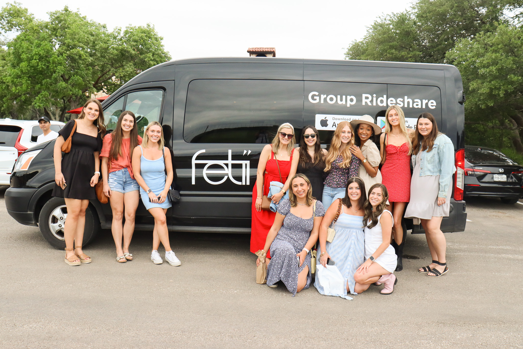 Group of ten women standing and sitting in front of a black van with text 'Group Rideshare' and 'feat' on it, outdoors.