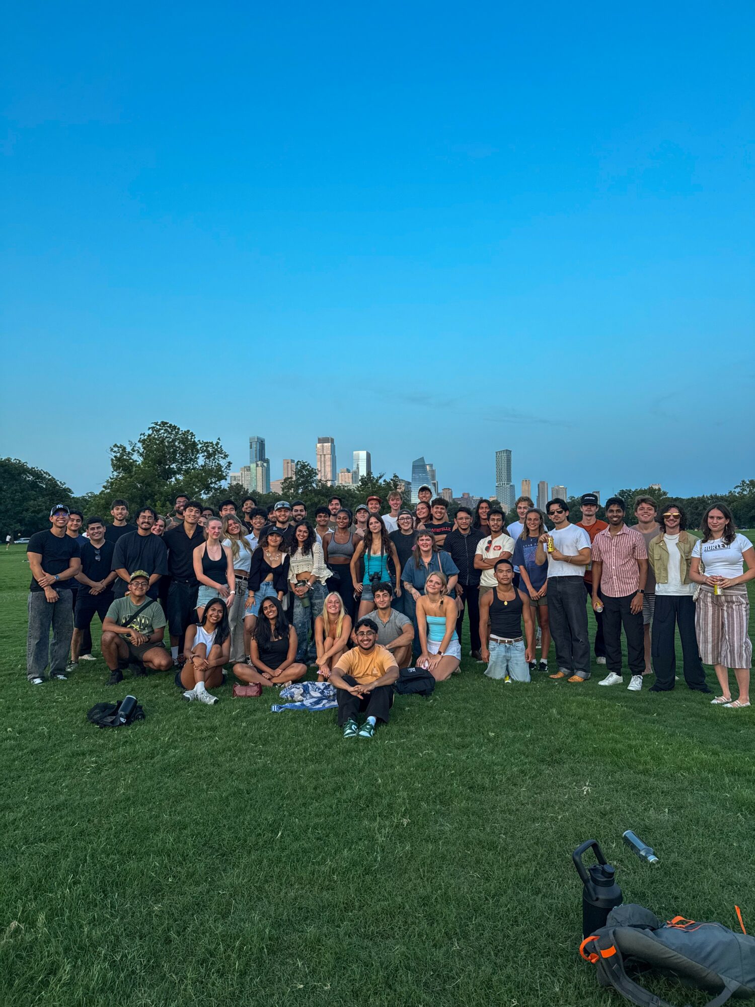 Group of people gathered outdoors on grass with city skyline in background, clear blue sky, some sitting and others standing.