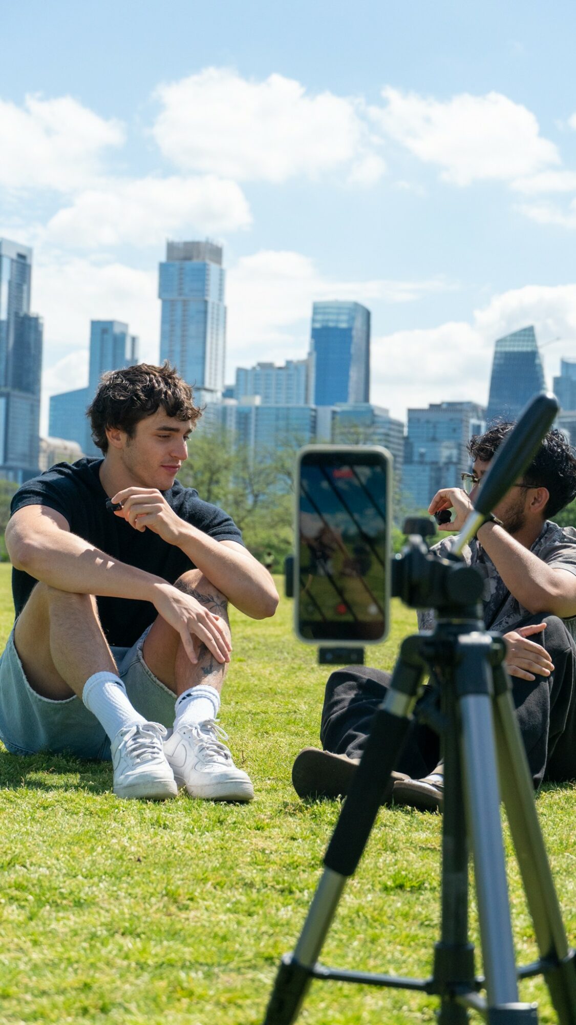Two people sitting on grass with city skyline in background, one holding a smartphone, camera on tripod nearby.