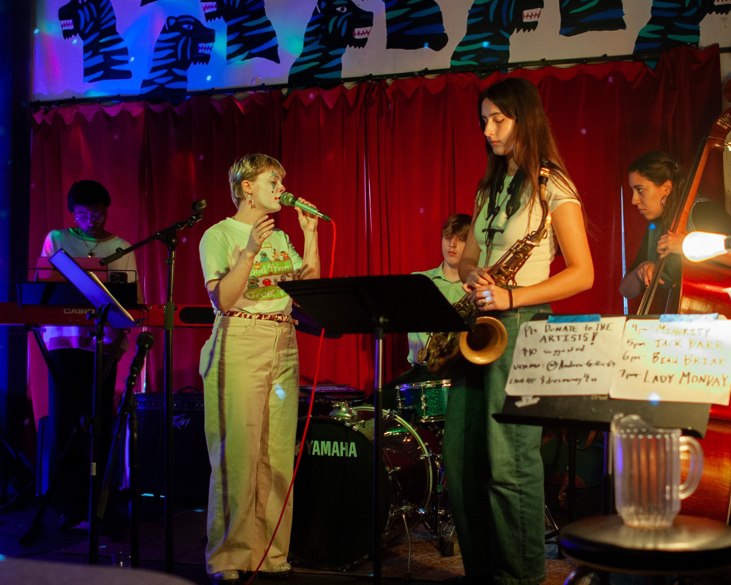 Group of children performing music on stage with instruments and microphone, red curtain backdrop, colorful lighting, and music stand.