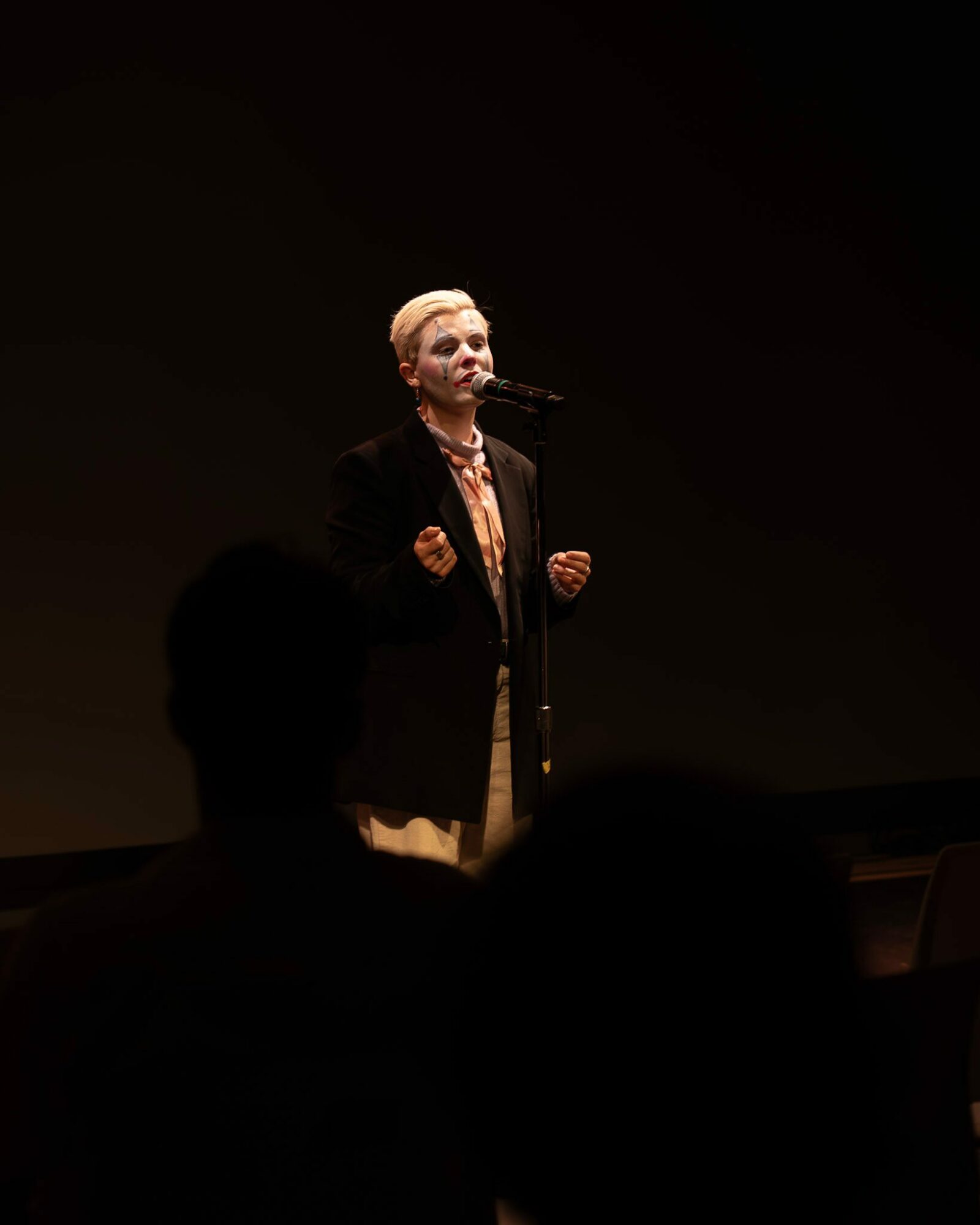 Woman speaking into a microphone on stage, with a dark background and an audience silhouette in foreground.