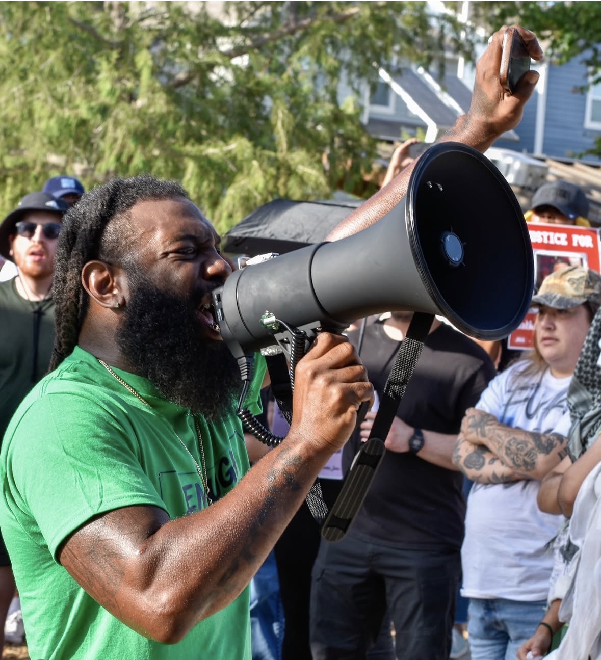 Man with a beard and green shirt speaking into a megaphone at outdoor gathering, surrounded by people and trees.