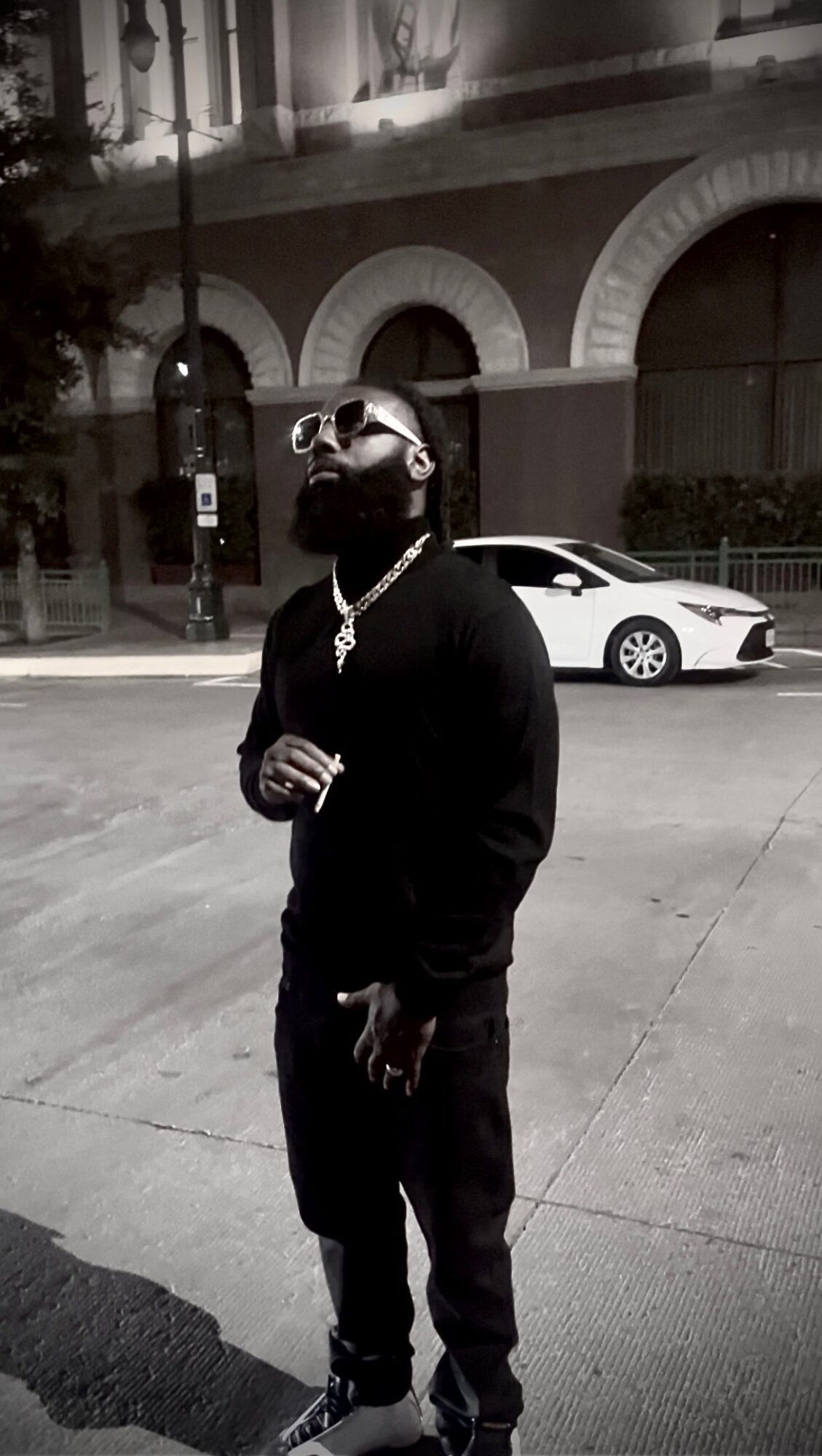Man with sunglasses and a chain stands on sidewalk at night, with a car and building in background.