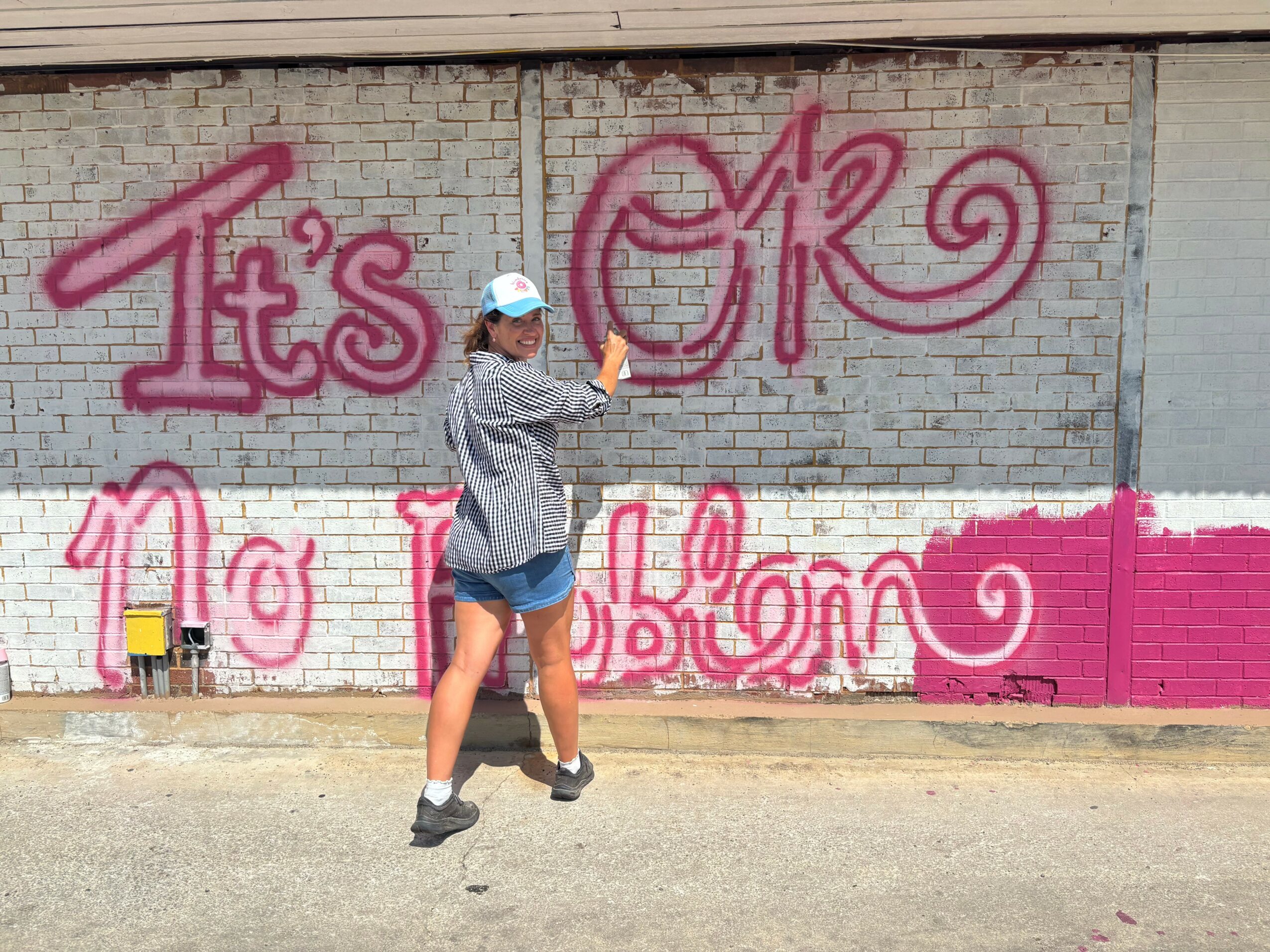 Person painting pink graffiti on a brick wall, wearing a hat and shorts, with a spray can in hand.