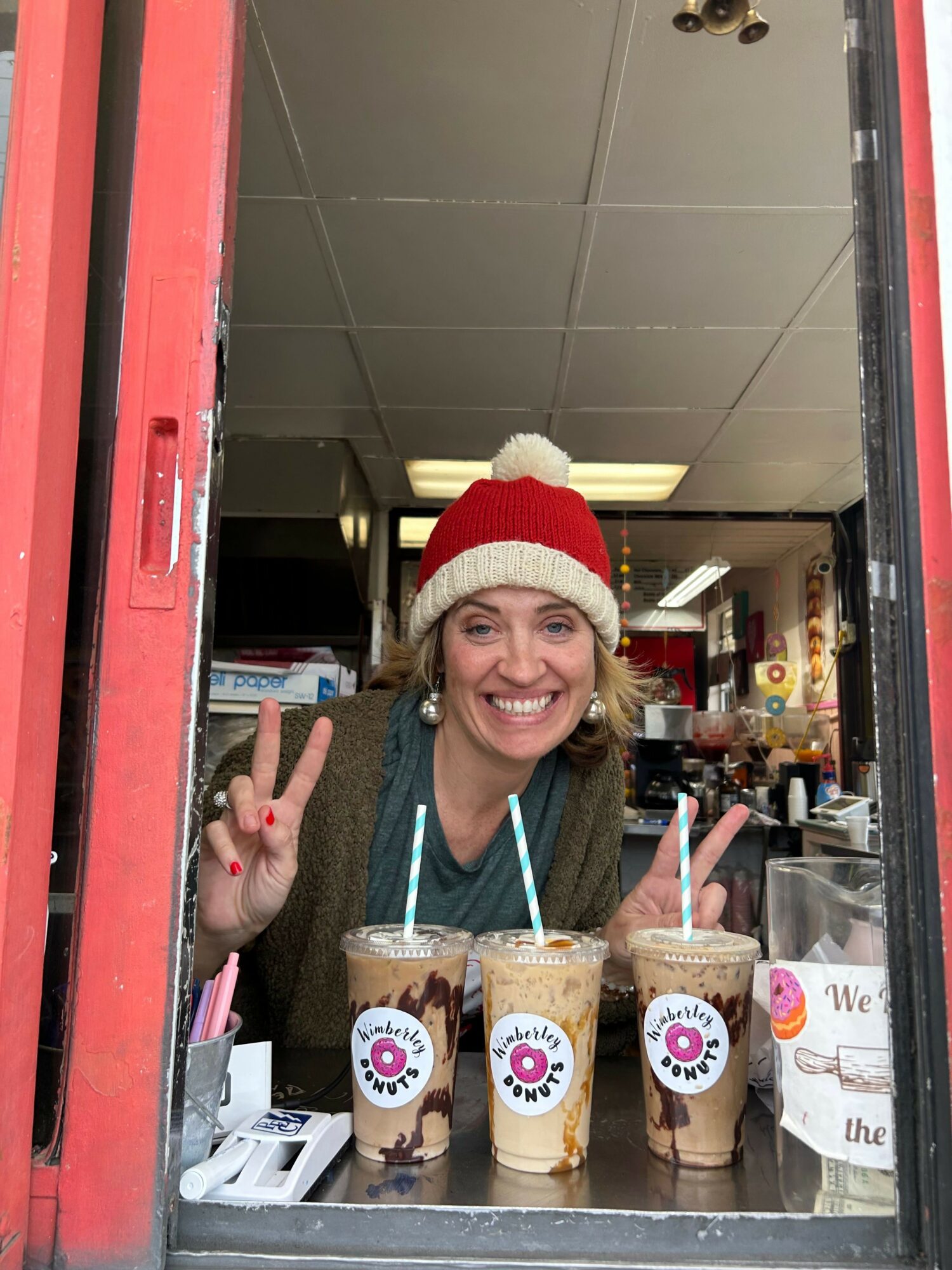 Person wearing a Santa hat smiling and making a peace sign at a counter with drinks, in a store or cafe.