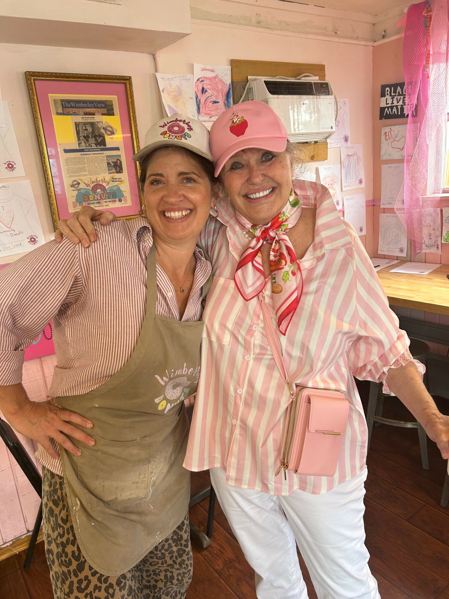 Two women smiling, one holding a framed certificate, in a room with pink and white decorations, one wearing a pink cap.