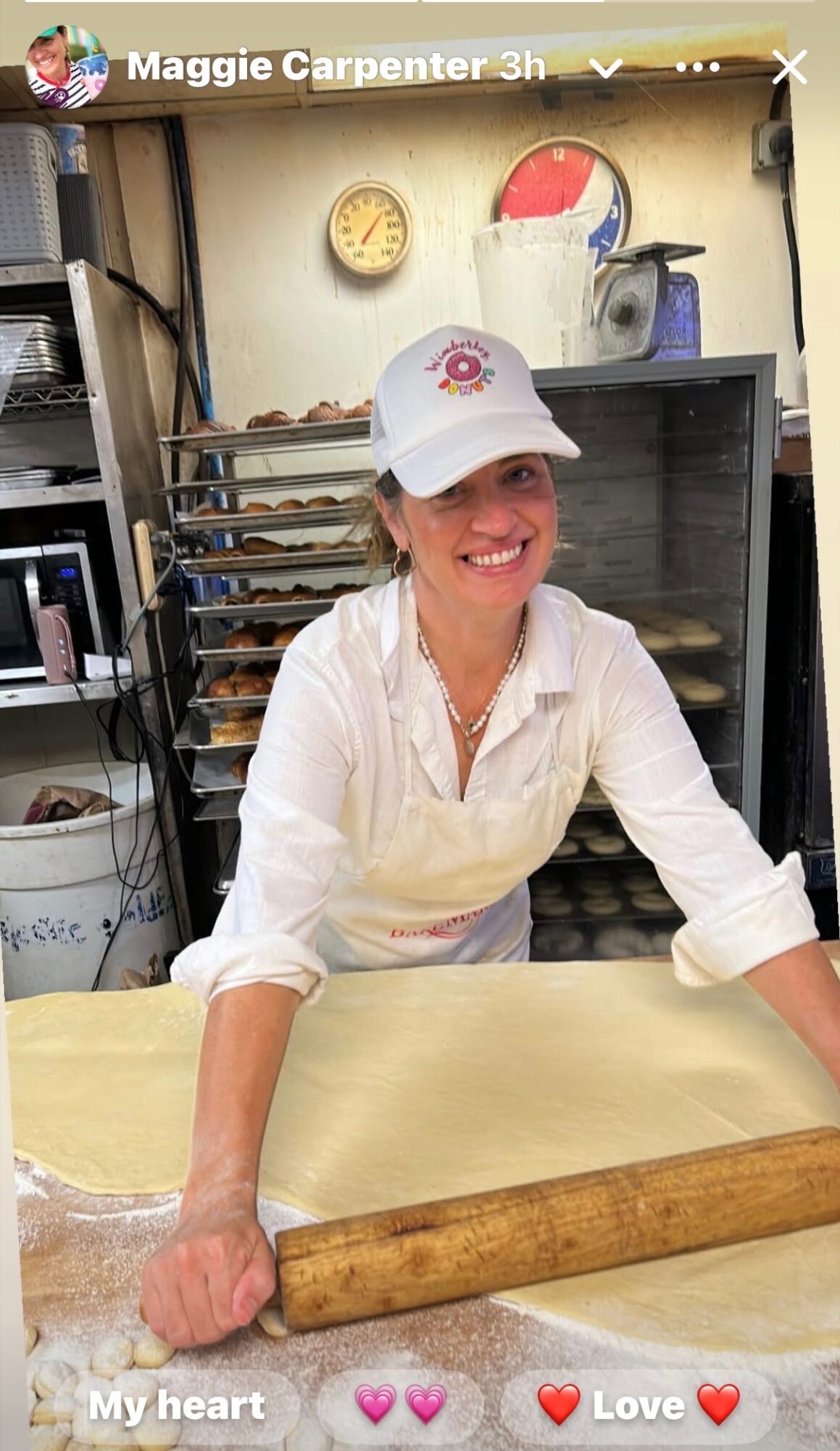 Woman wearing a white cap and apron smiling at a bakery counter with a rolling pin.