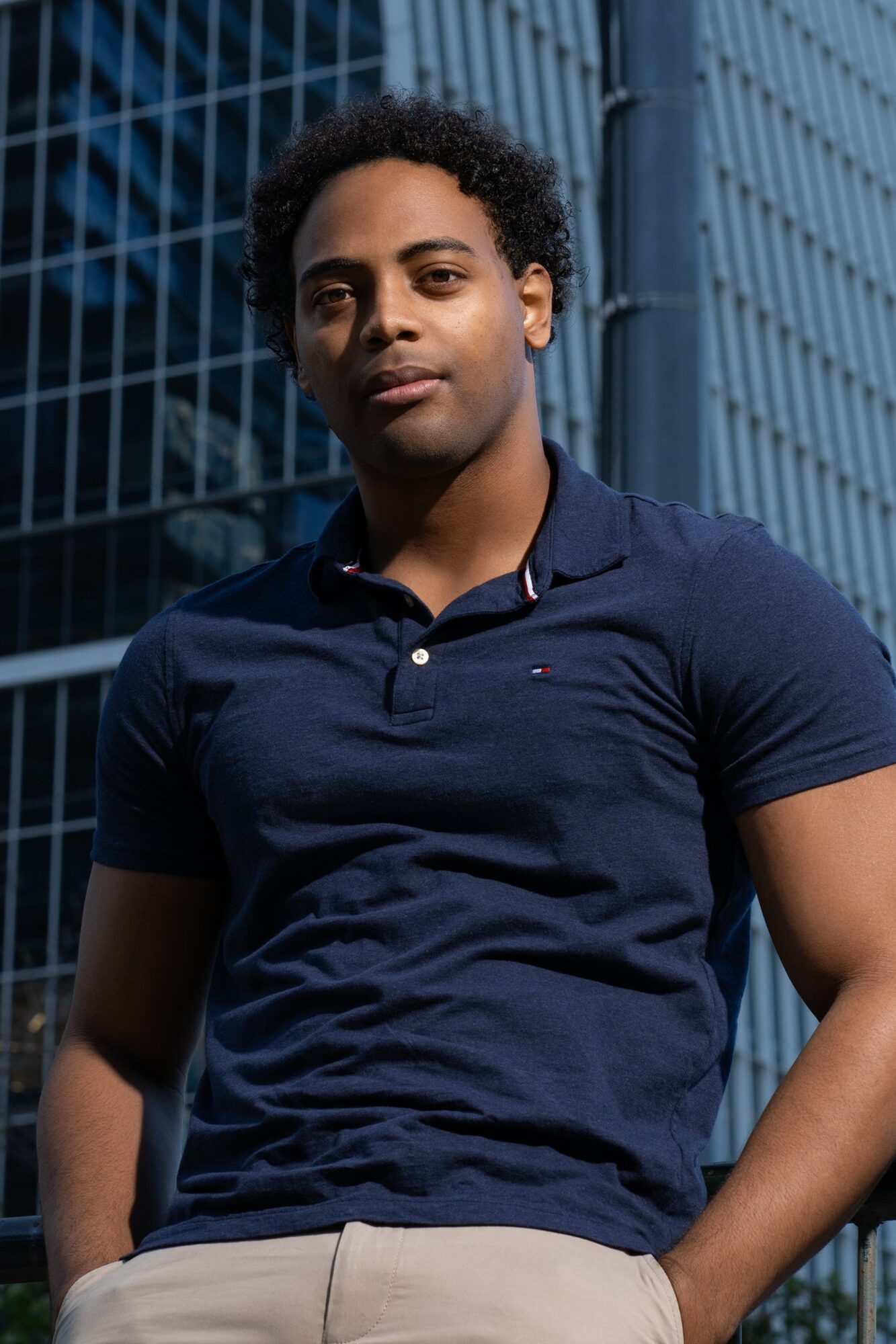 Young man with curly hair wearing a navy polo shirt and beige pants standing outdoors in front of a modern building.