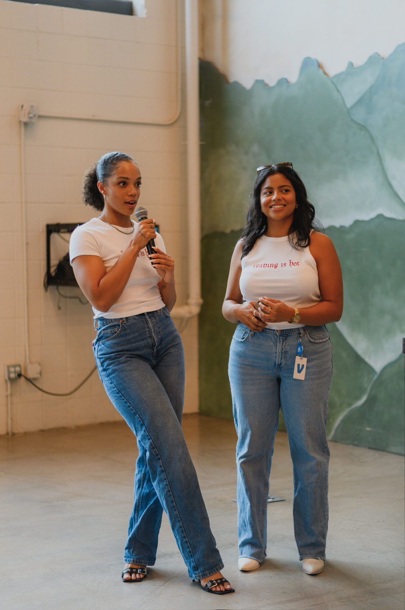 Two women standing indoors, one speaking into a microphone, the other smiling, with a painted mountain mural behind them.