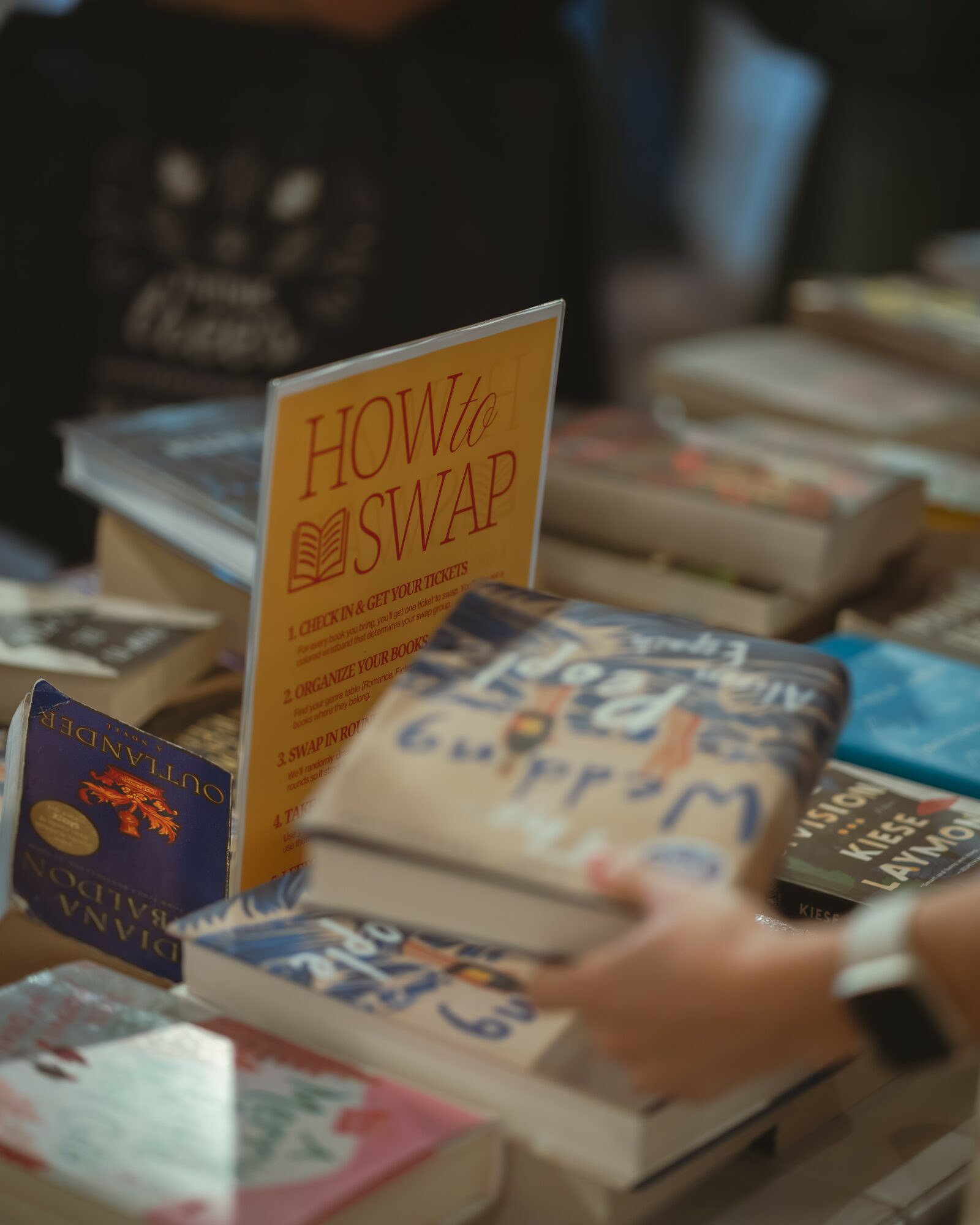 Person holding a book at a table with various books and a yellow flyer titled 'How to Swap'.