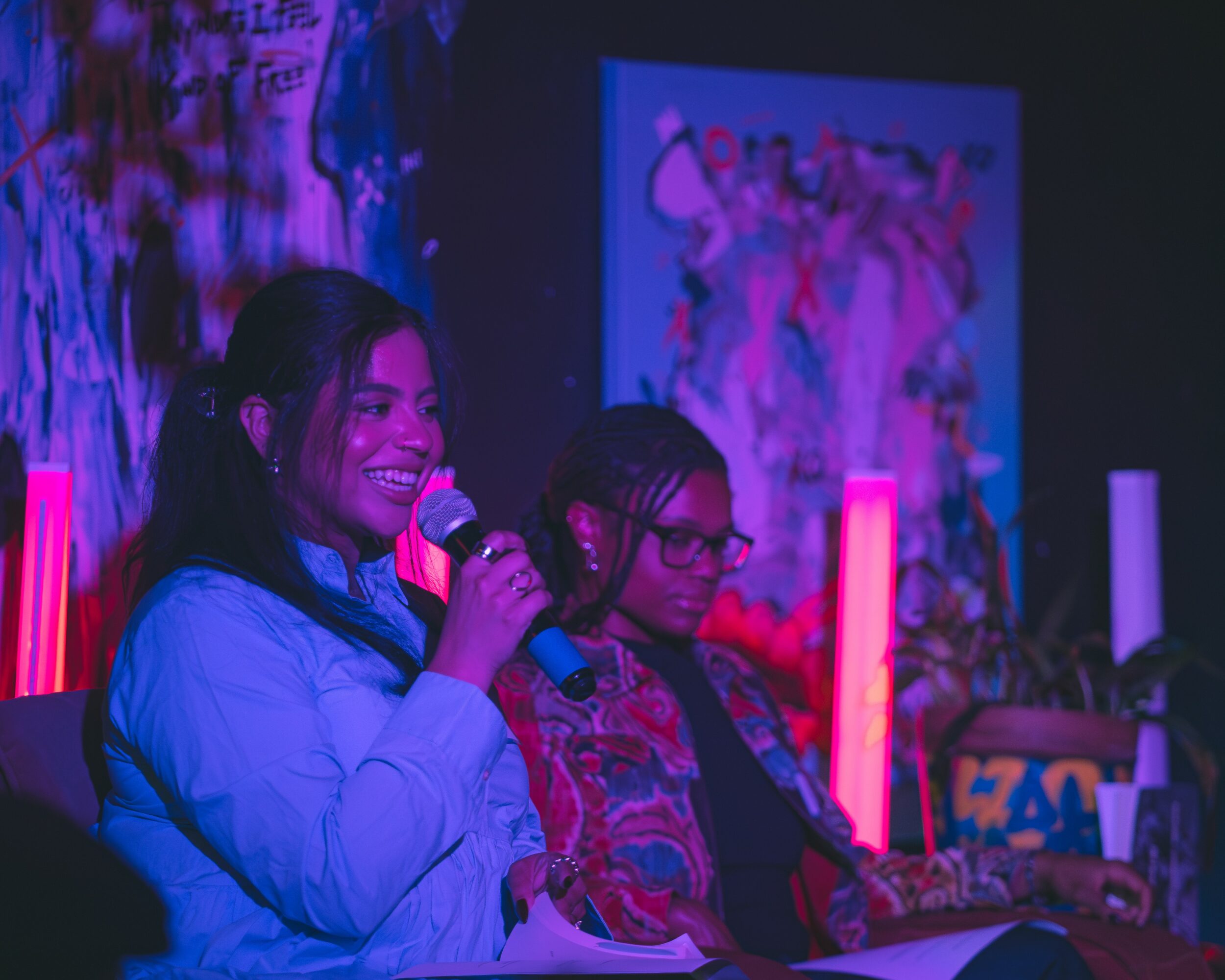 Two women sit on a stage, one smiling and holding a microphone, with colorful lights and artwork in the background.