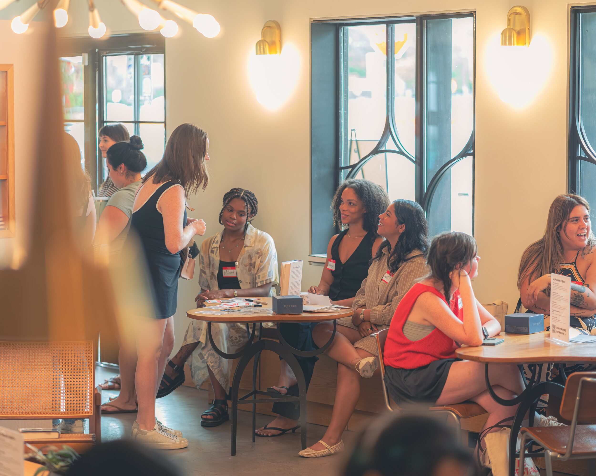 Group of women sitting at tables in a well-lit cafe, engaging in conversation, with large windows in the background.