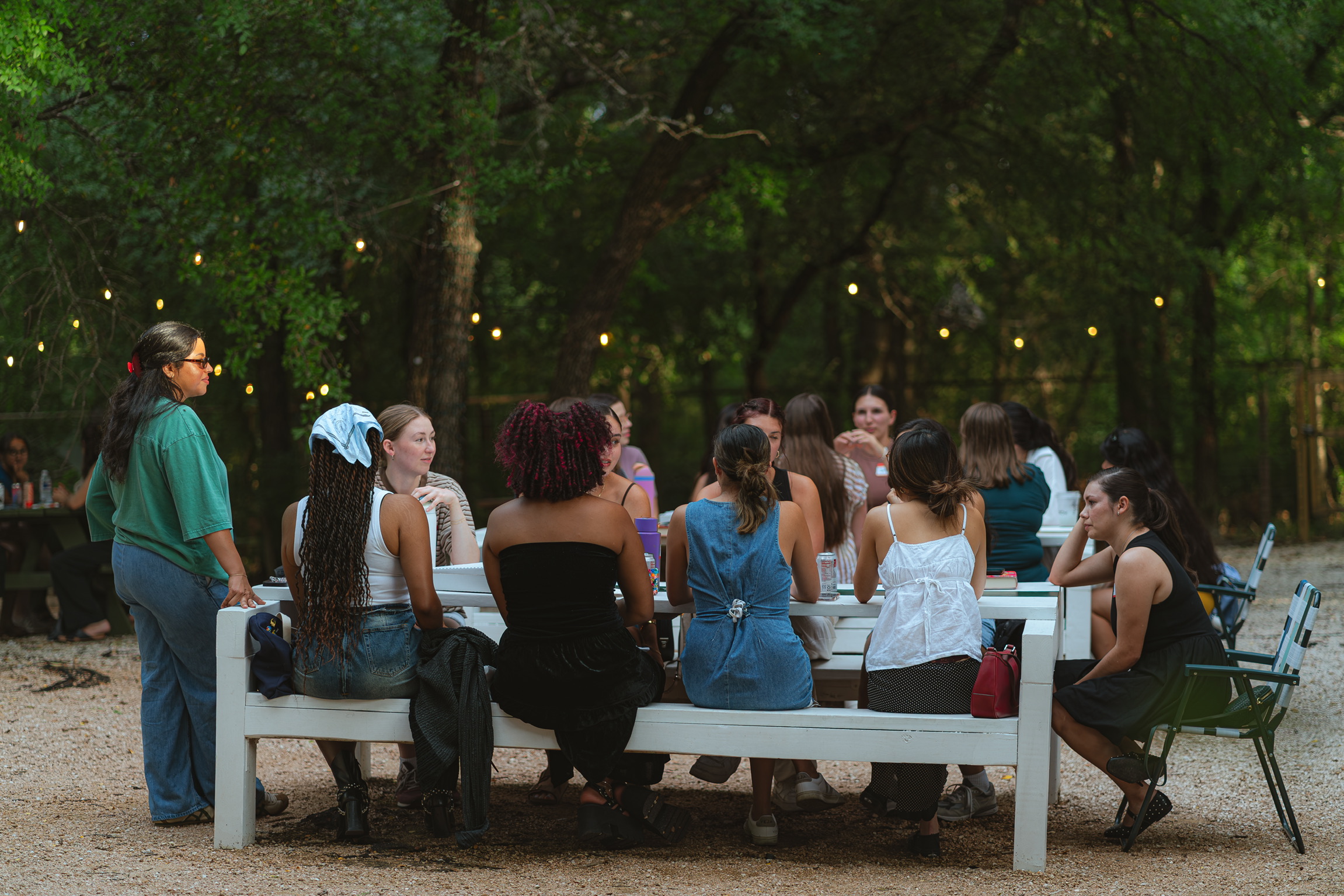 Group of people sitting on a bench outdoors, surrounded by trees and string lights, engaged in conversation.