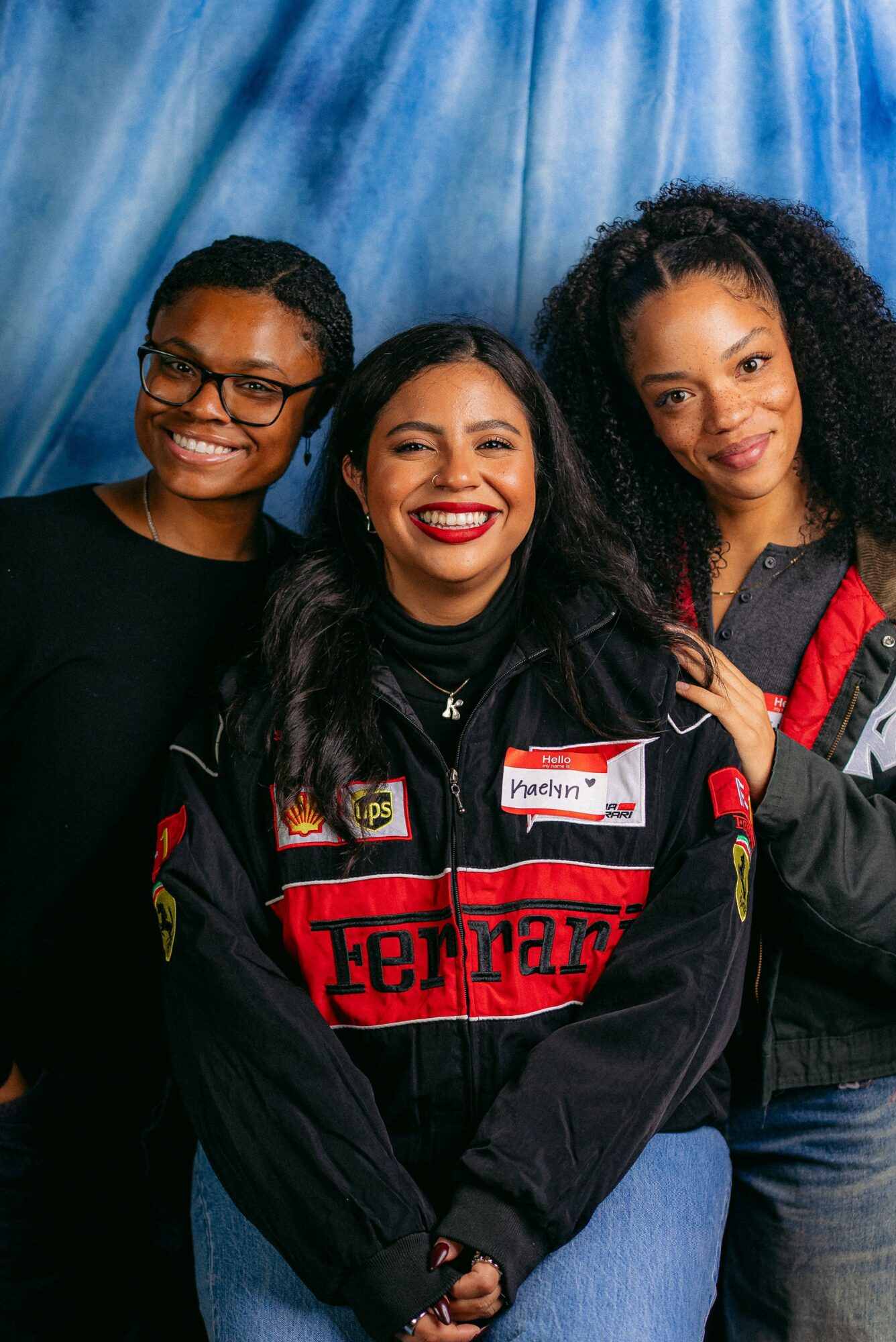 Three women smiling, standing close together, with a blue curtain background.