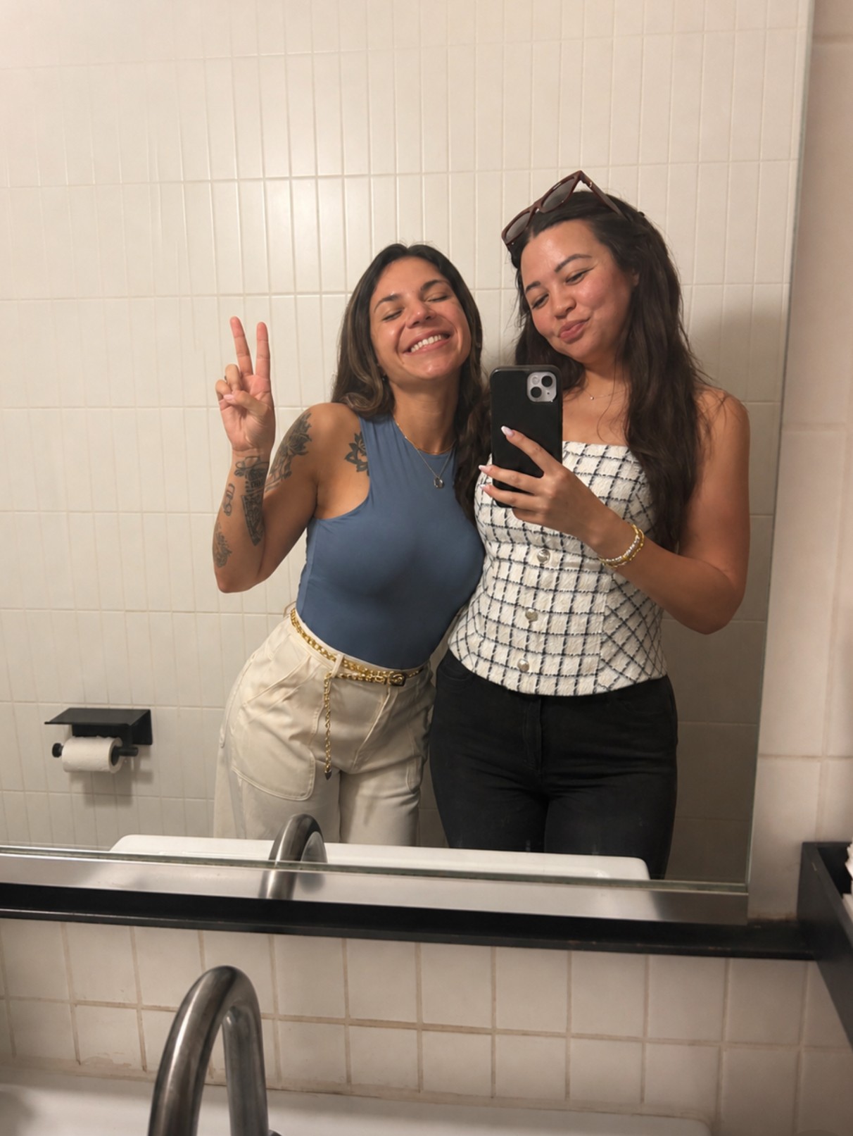 Two women taking a mirror selfie in a bathroom, smiling, one making a peace sign, with white tiled walls.