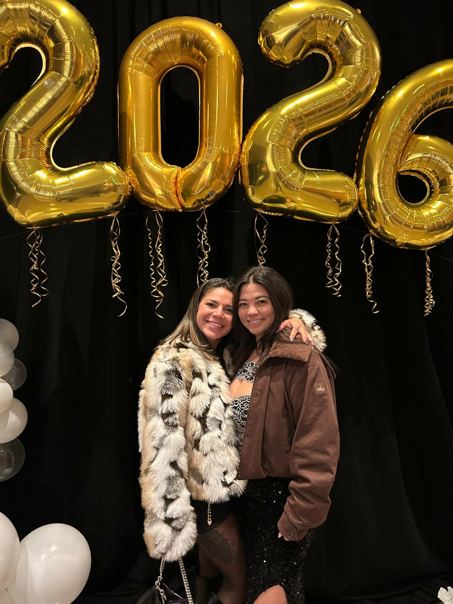Two women standing close together, smiling, with large gold balloons spelling '2023' and white balloons behind them.