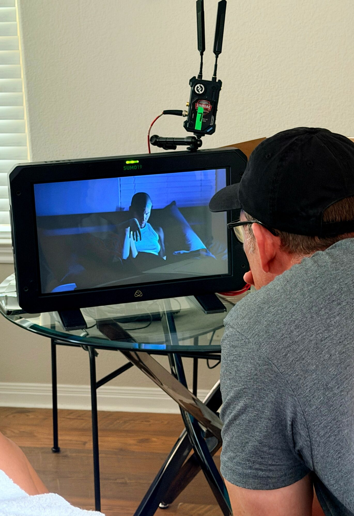 Person watching a video on a monitor, with a camera mounted above, in a room with a window and wooden floor.