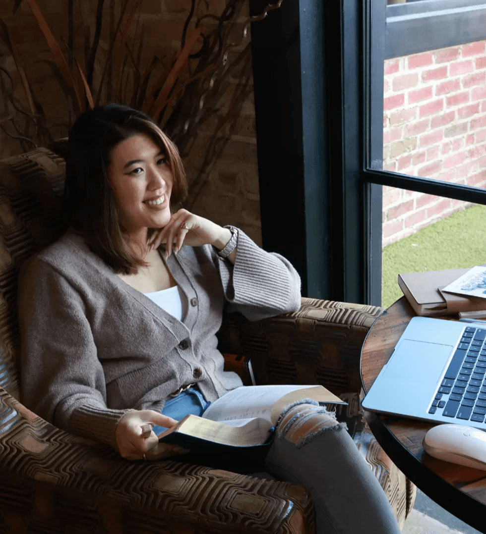 Woman sitting in a cozy room near a window, reading a book, with a laptop on the table beside her.