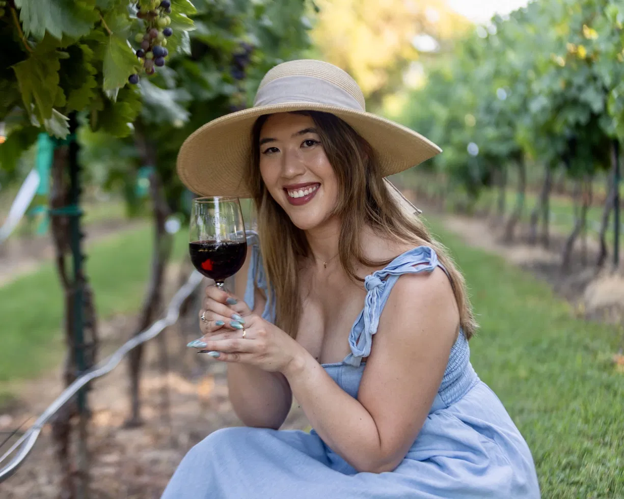 Woman in a blue dress and wide-brimmed hat smiling and holding a glass of red wine outdoors in a vineyard.