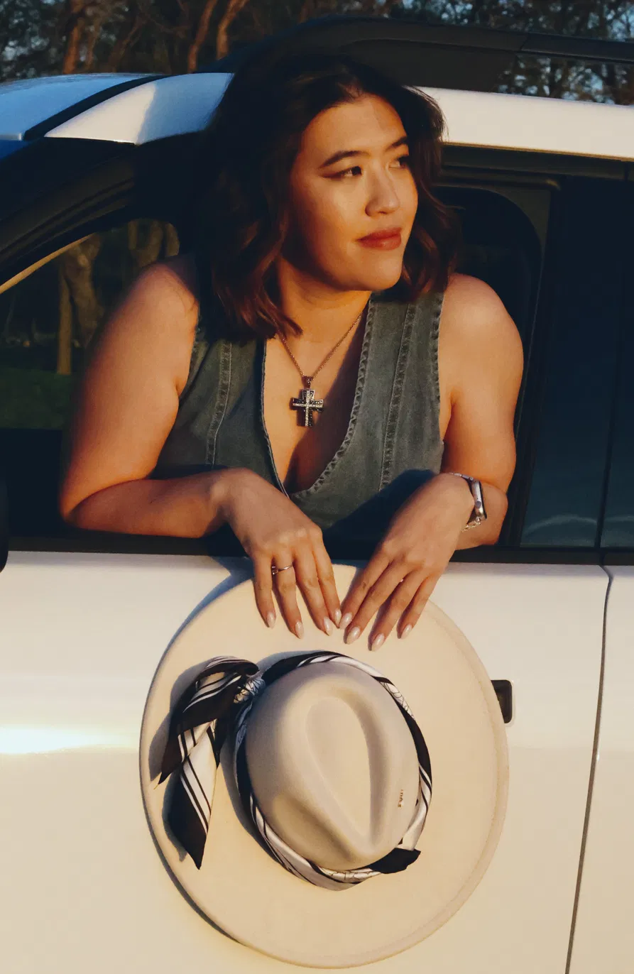 Woman with wavy hair leaning on car window, wearing a sleeveless top and cross necklace, holding a hat.