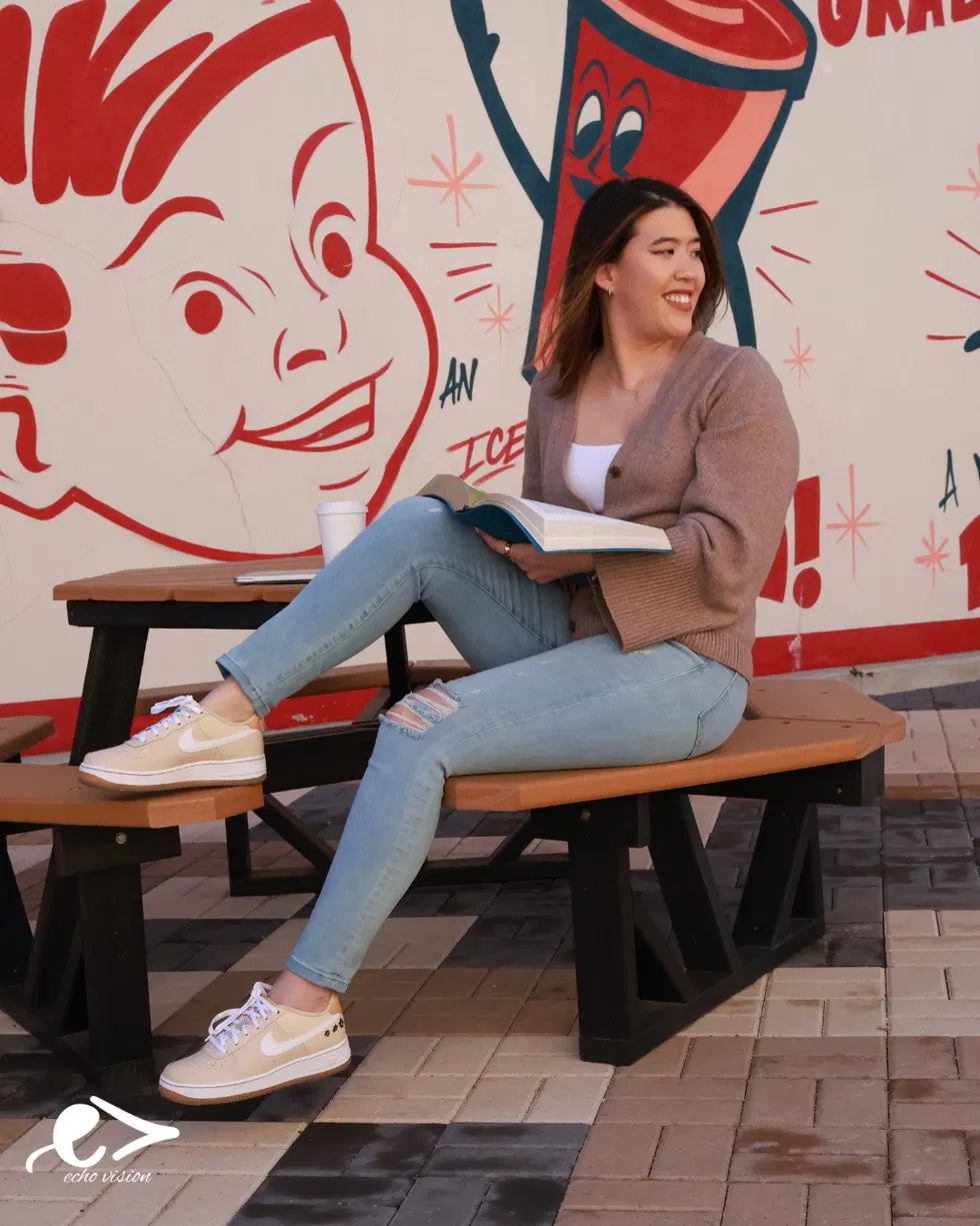 Young woman sitting on a bench, holding a book, smiling, with a colorful mural background featuring a cartoon drink and face.