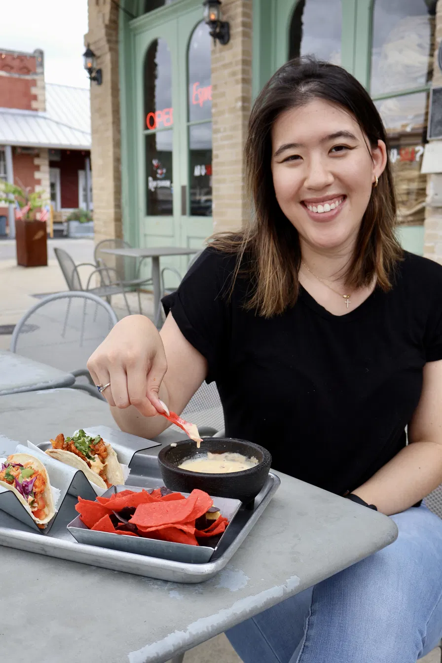Smiling woman sitting at outdoor table with food, in front of a green storefront, wearing a black shirt and jeans.