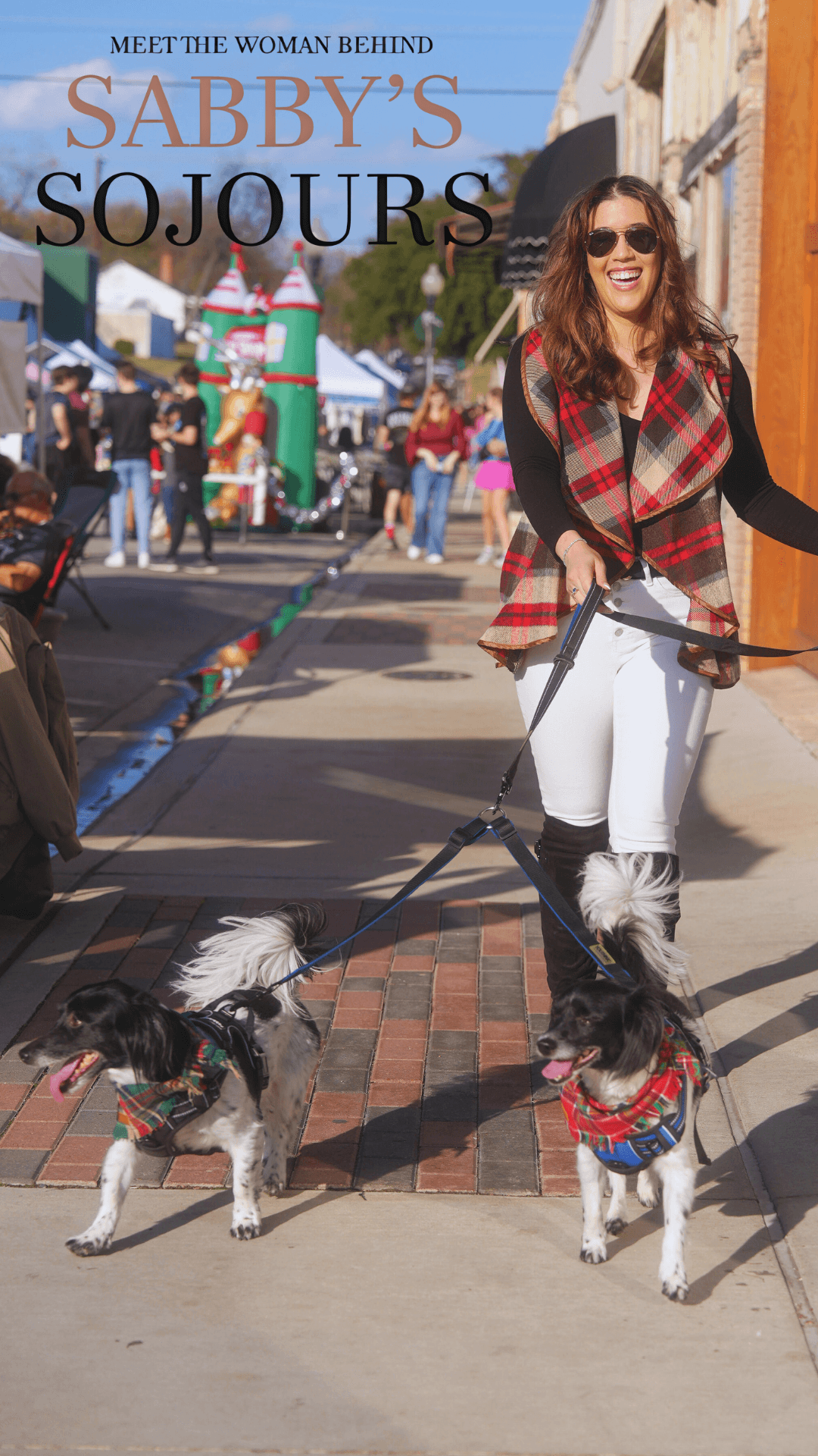Woman walking two dogs on a city street, smiling, wearing sunglasses, a plaid vest, and white pants.