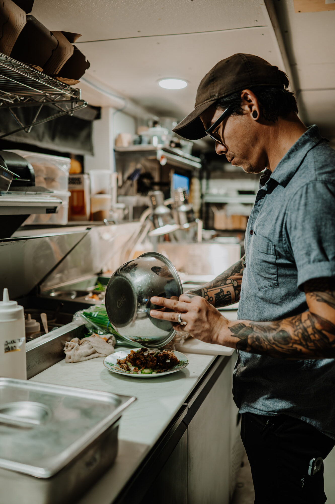 Person with tattoos and glasses pouring food onto a plate in a kitchen, with shelves and utensils in the background.