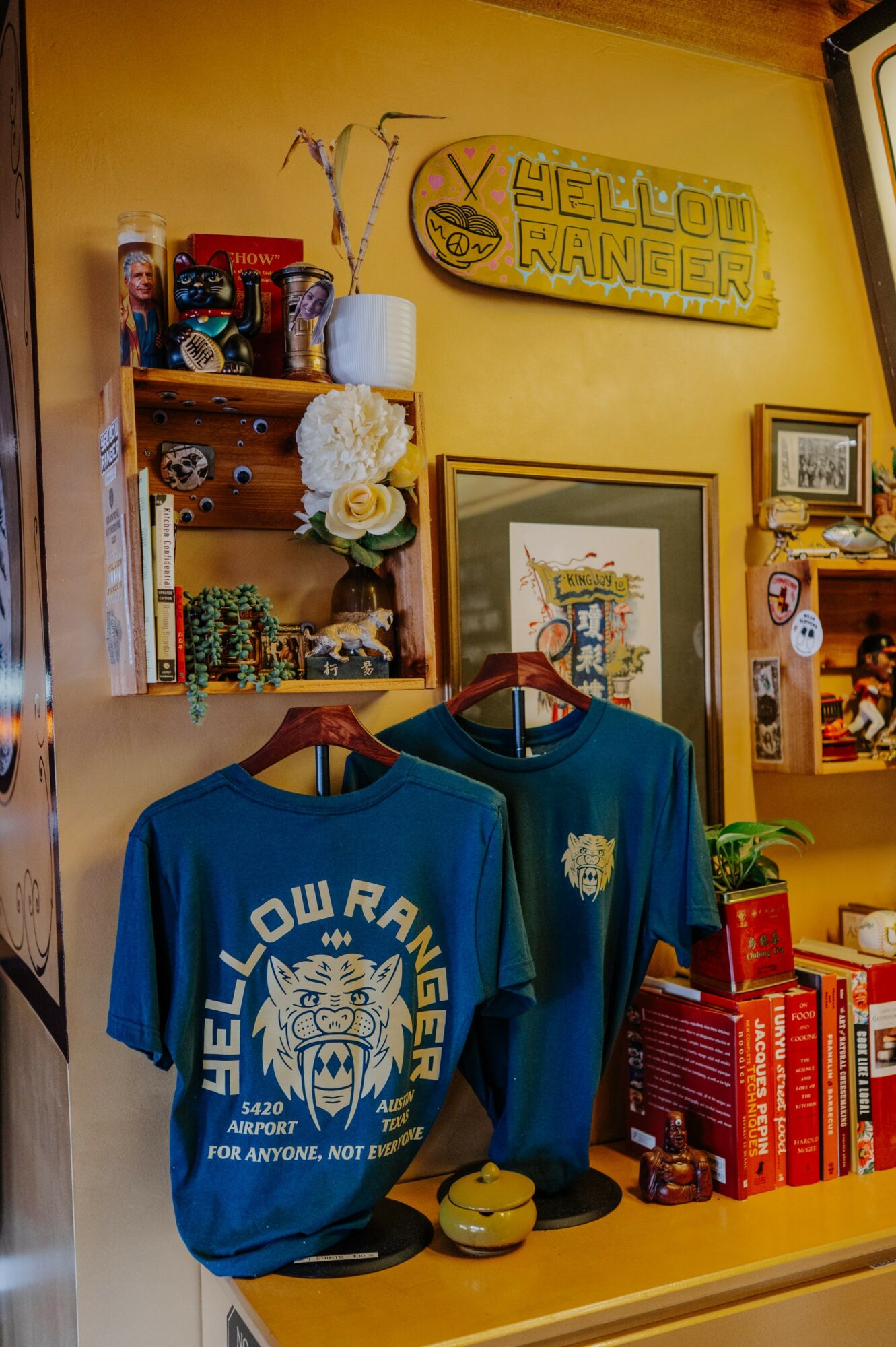 Two blue T-shirts with yellow and white designs hang on a wooden shelf, surrounded by books, framed pictures, and decorative items.