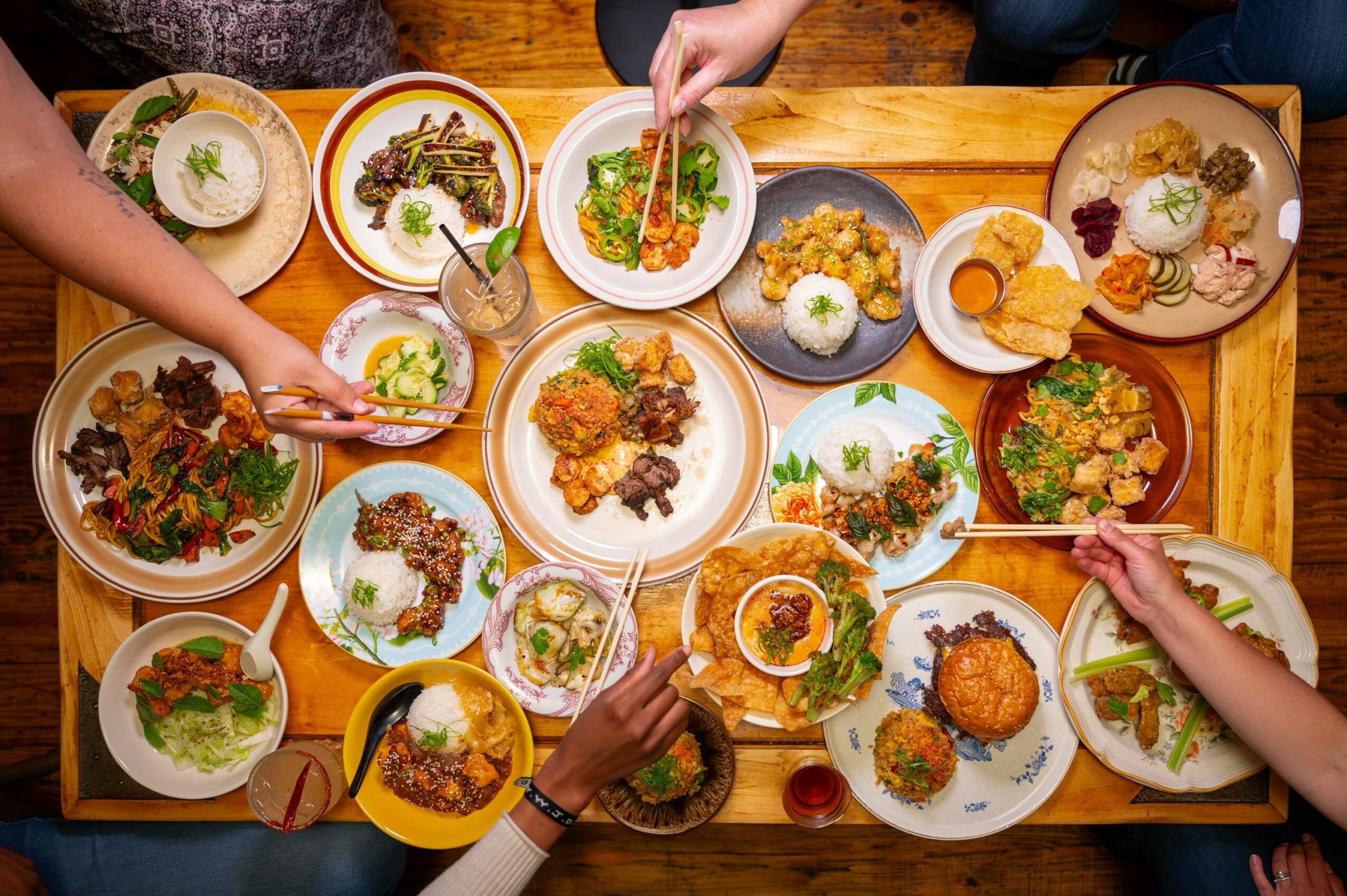 People sharing a variety of dishes on a wooden table, including salads, rice, and fried items, with hands reaching for food.