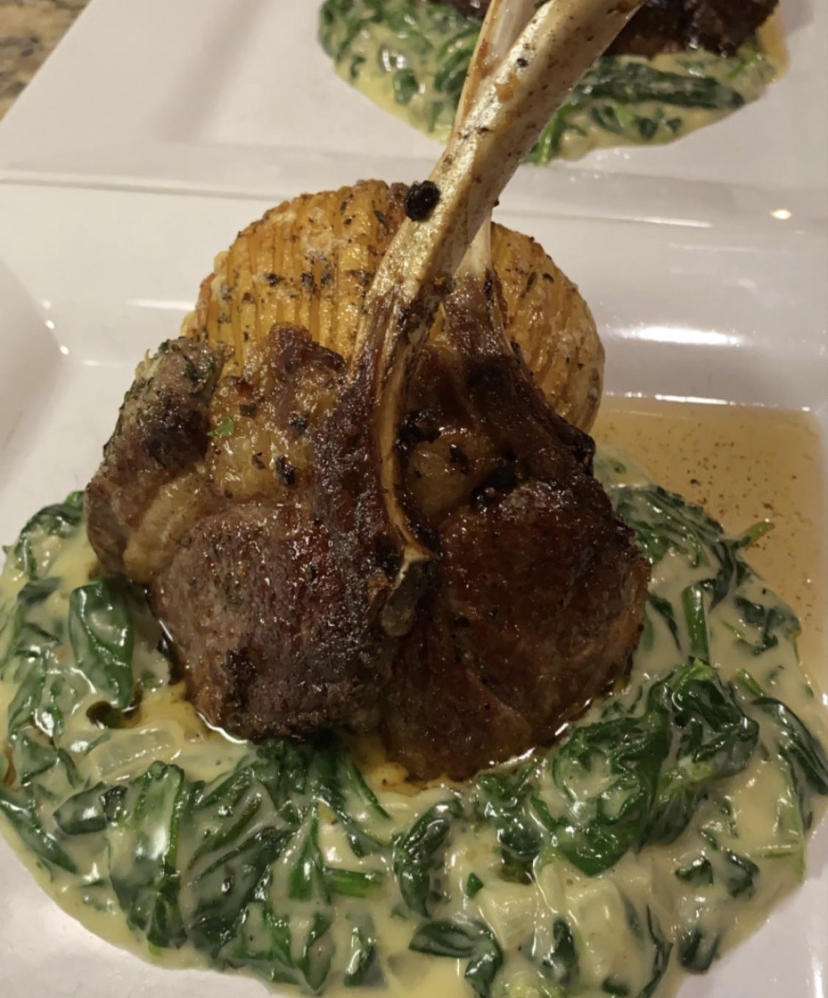 A plated dish with a cooked meat chop, mashed potatoes, and green leafy vegetables in sauce.