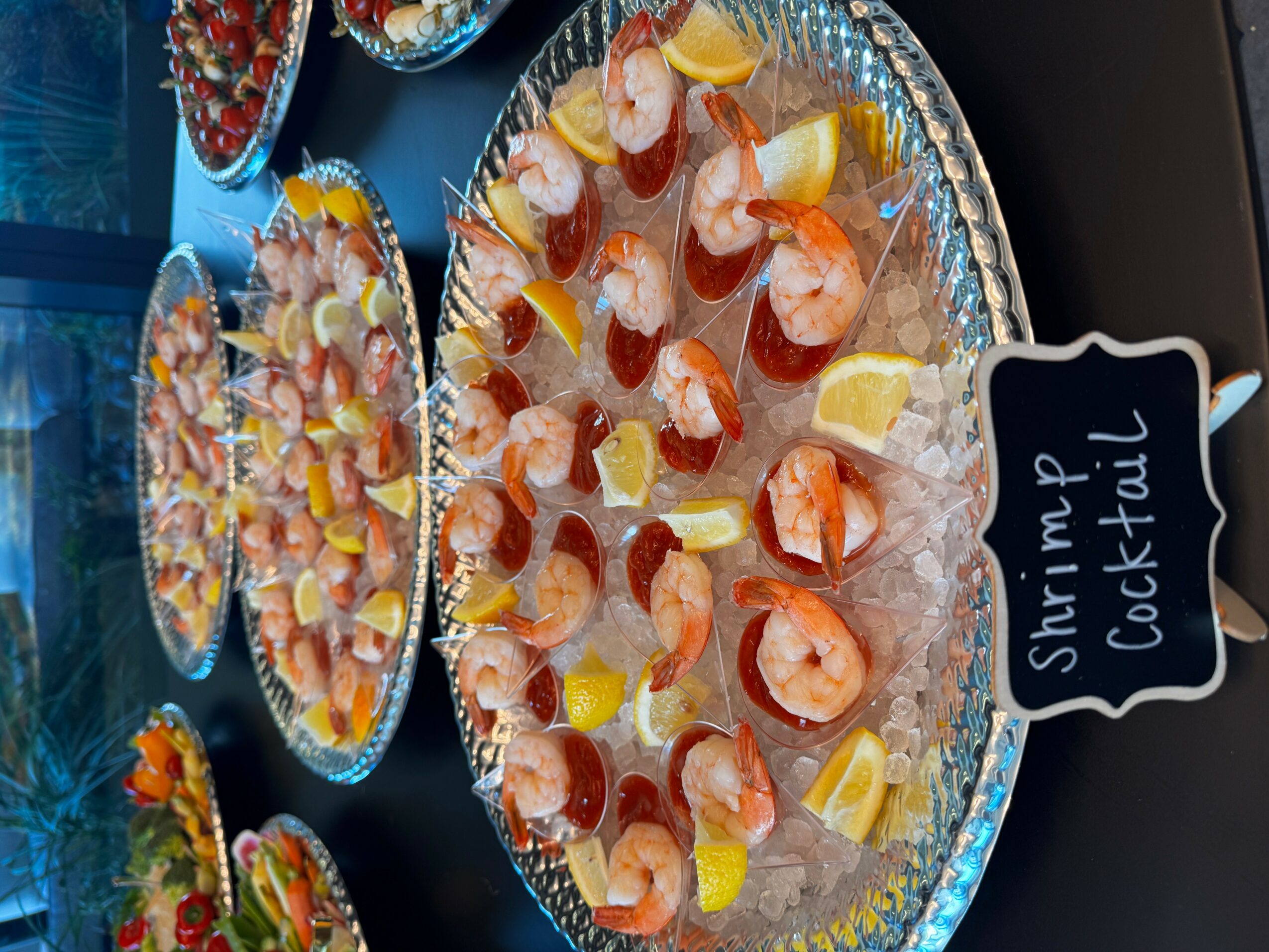 Assorted shrimp cocktail appetizers with lemon wedges on a tray, labeled 'Shrimp Cocktail' in a small sign, viewed from above.