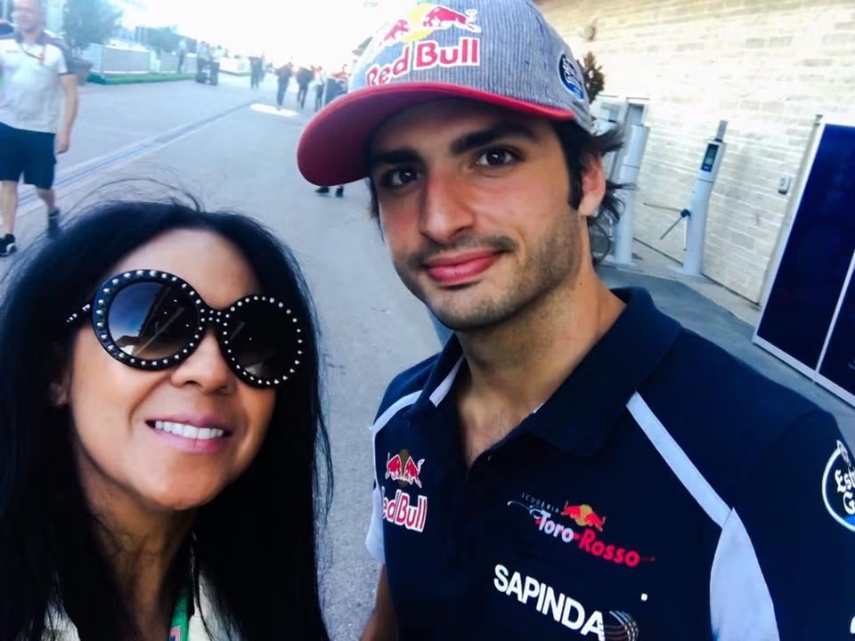 Two people smiling outdoors, one wearing large sunglasses and the other in a racing team shirt and cap.