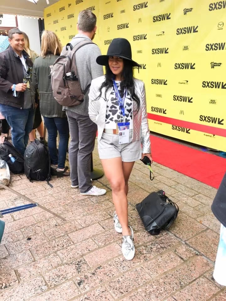 Woman in a black hat, white shorts, and patterned shirt standing on a red carpet at SXSW event, with people and yellow SXSW backdrop behind her.