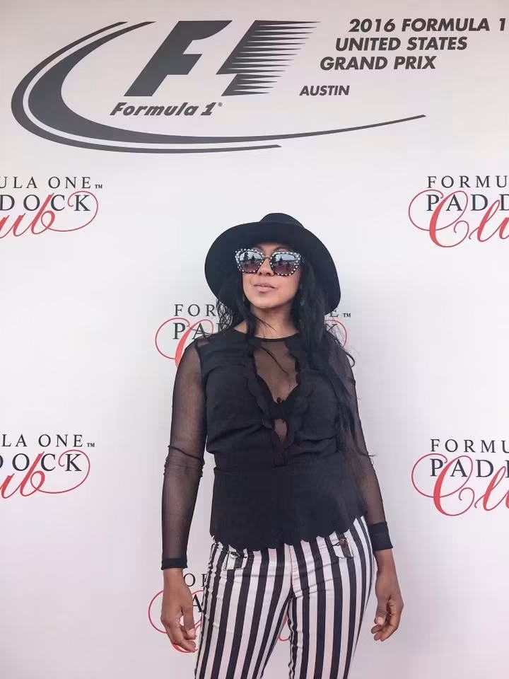 Woman in sunglasses and black outfit standing in front of a Formula 1 backdrop, striped pants, black hat, long dark hair.