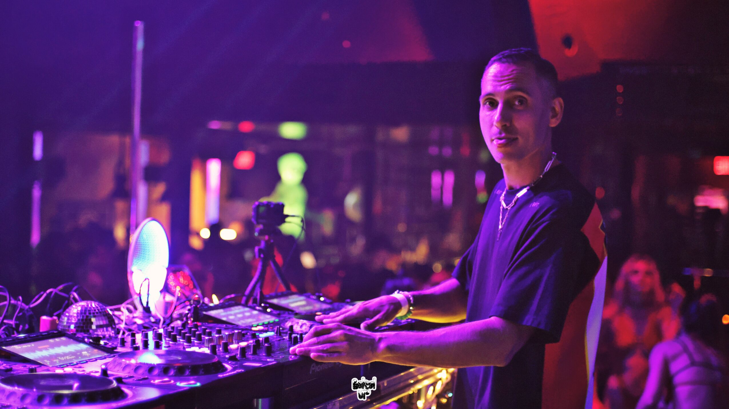 DJ with equipment in a colorful nightclub, people dancing in the background, vibrant lighting, and a focused expression.